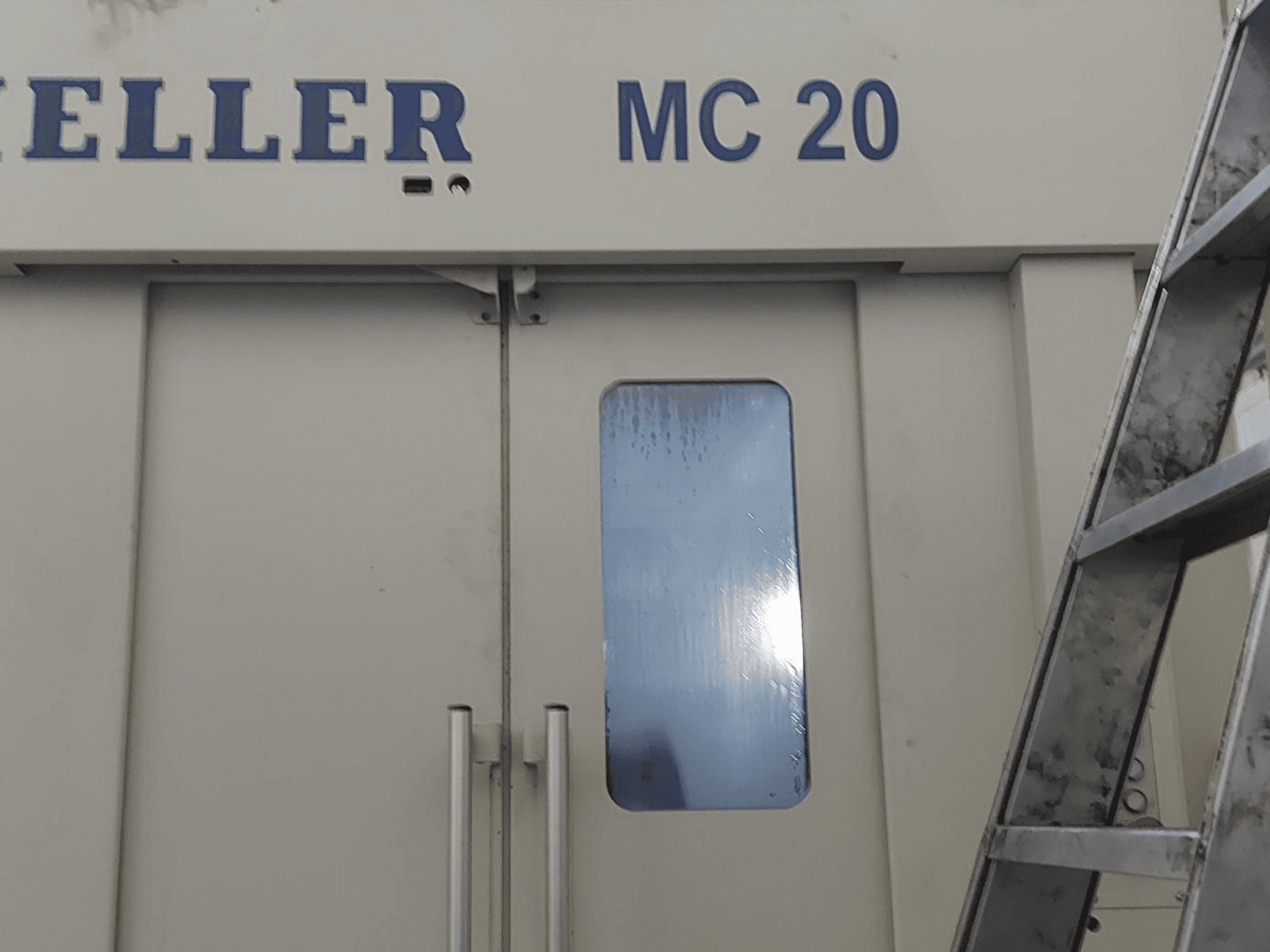 Vooraanzicht van HELLER MC 20 horizontaal bewerkingscentrum, met deurgrepen en een klein venster. Ladder aan de zijkant.