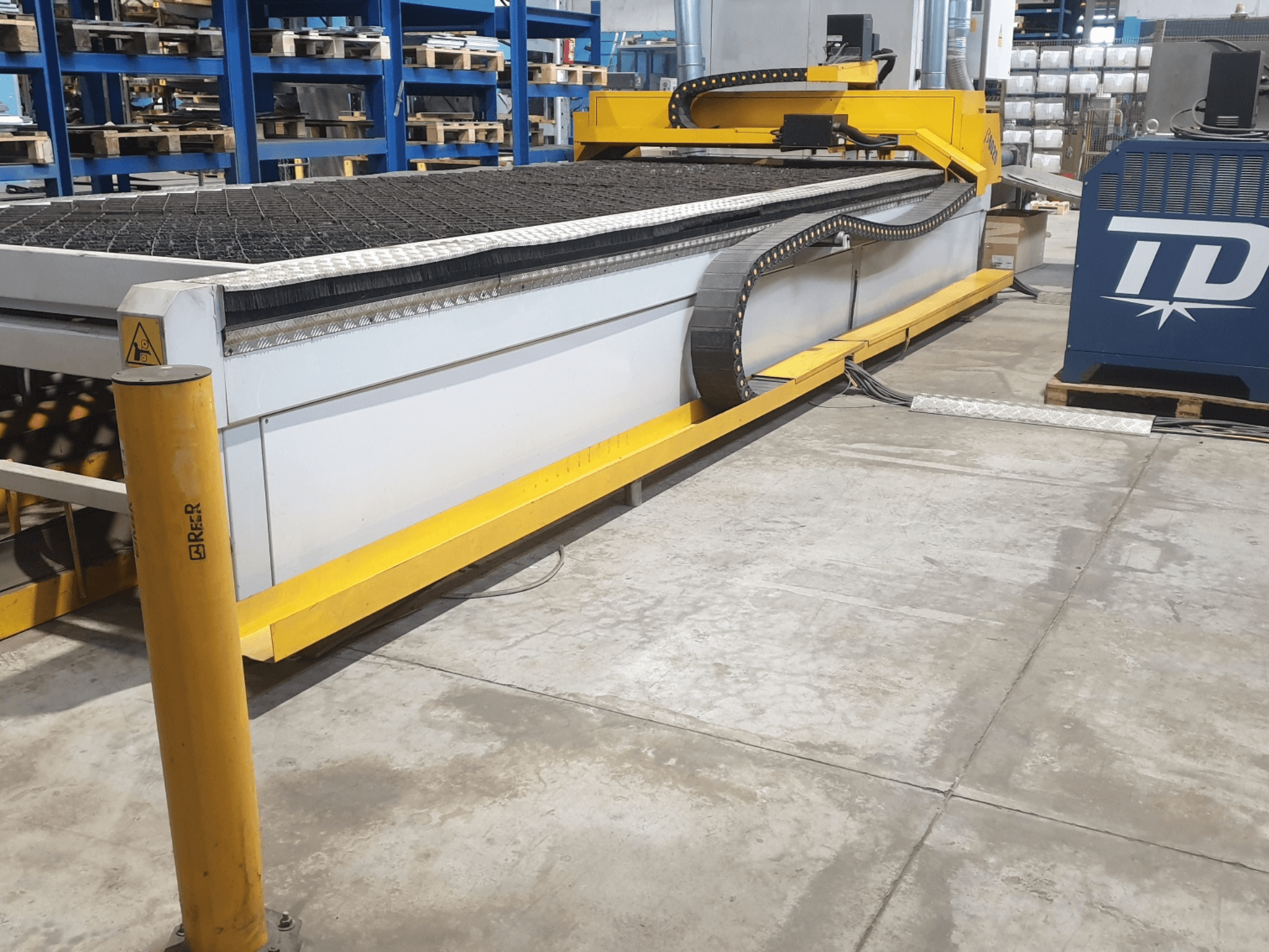 Industriële snijmachine met gele rand, vooraanzicht, met een gestructureerd bed en bedieningspaneel, geplaatst op een fabrieksvloer.