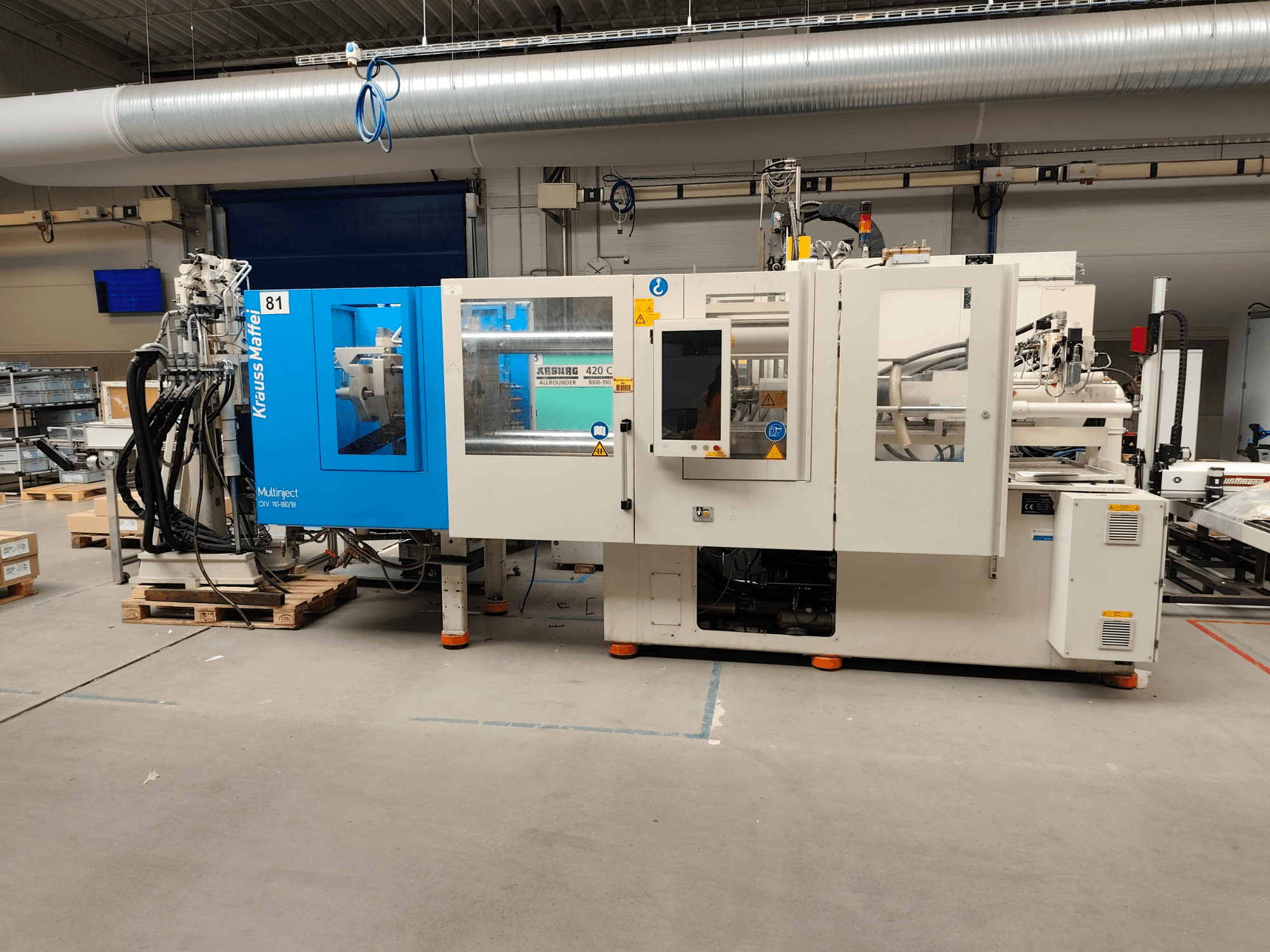 Krauss Maffei 110-180-180 CX V hydraulische spuitgietmachine, zijaanzicht, met blauw-witte behuizing, bedieningspaneel en aansluitingen.
