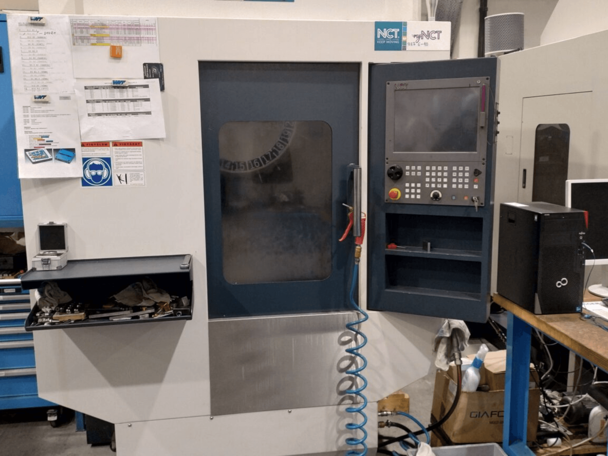 NCT CNC machine, vooraanzicht, voorzien van bedieningspaneel met numeriek toetsenbord, gereedschap in opberglade en handleiding aan de muur.