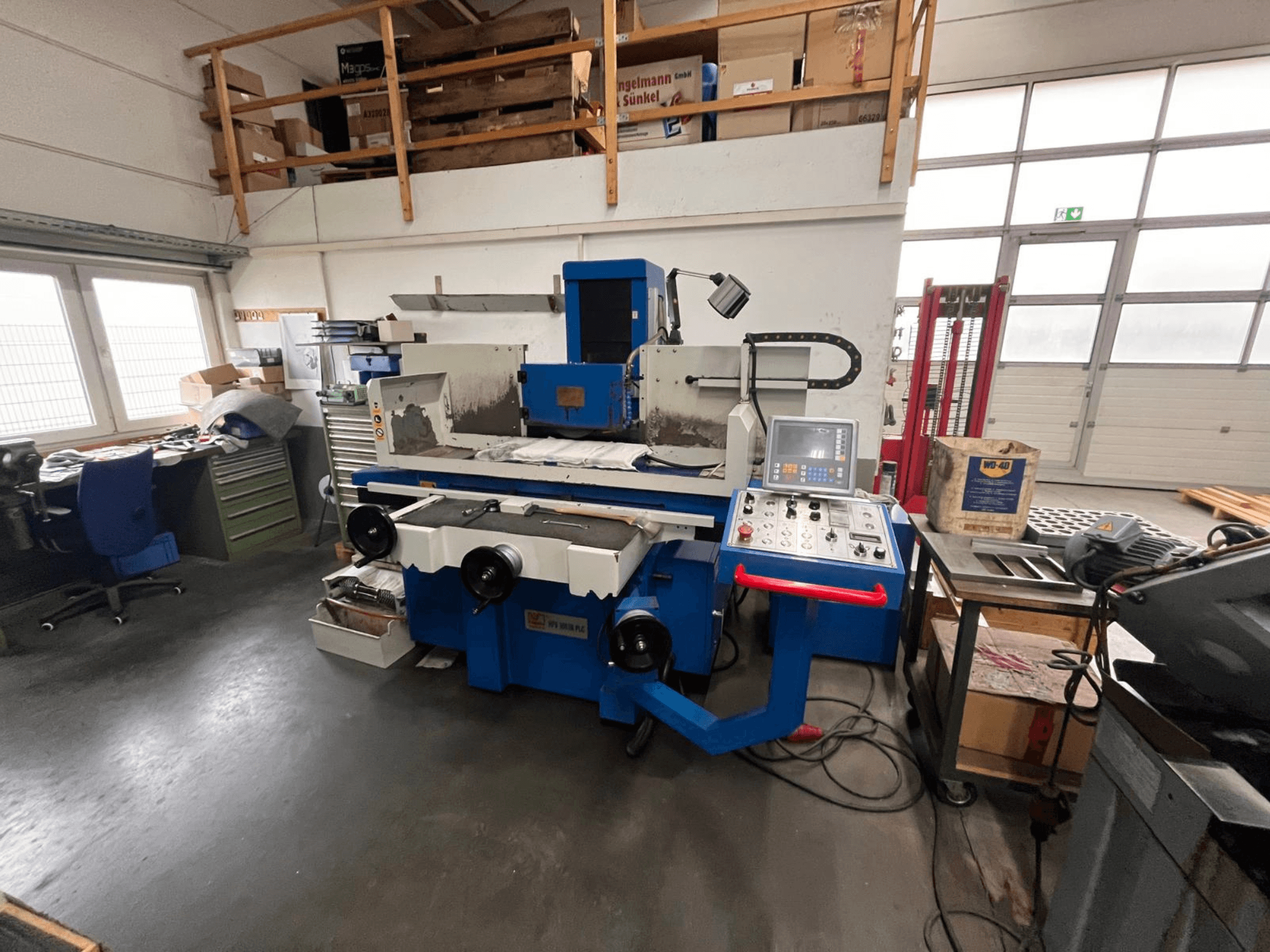 Blauwe vlakslijpmachine met digitaal bedieningspaneel, vooraanzicht, met een rommelige werkruimte en een groene gereedschapskast.