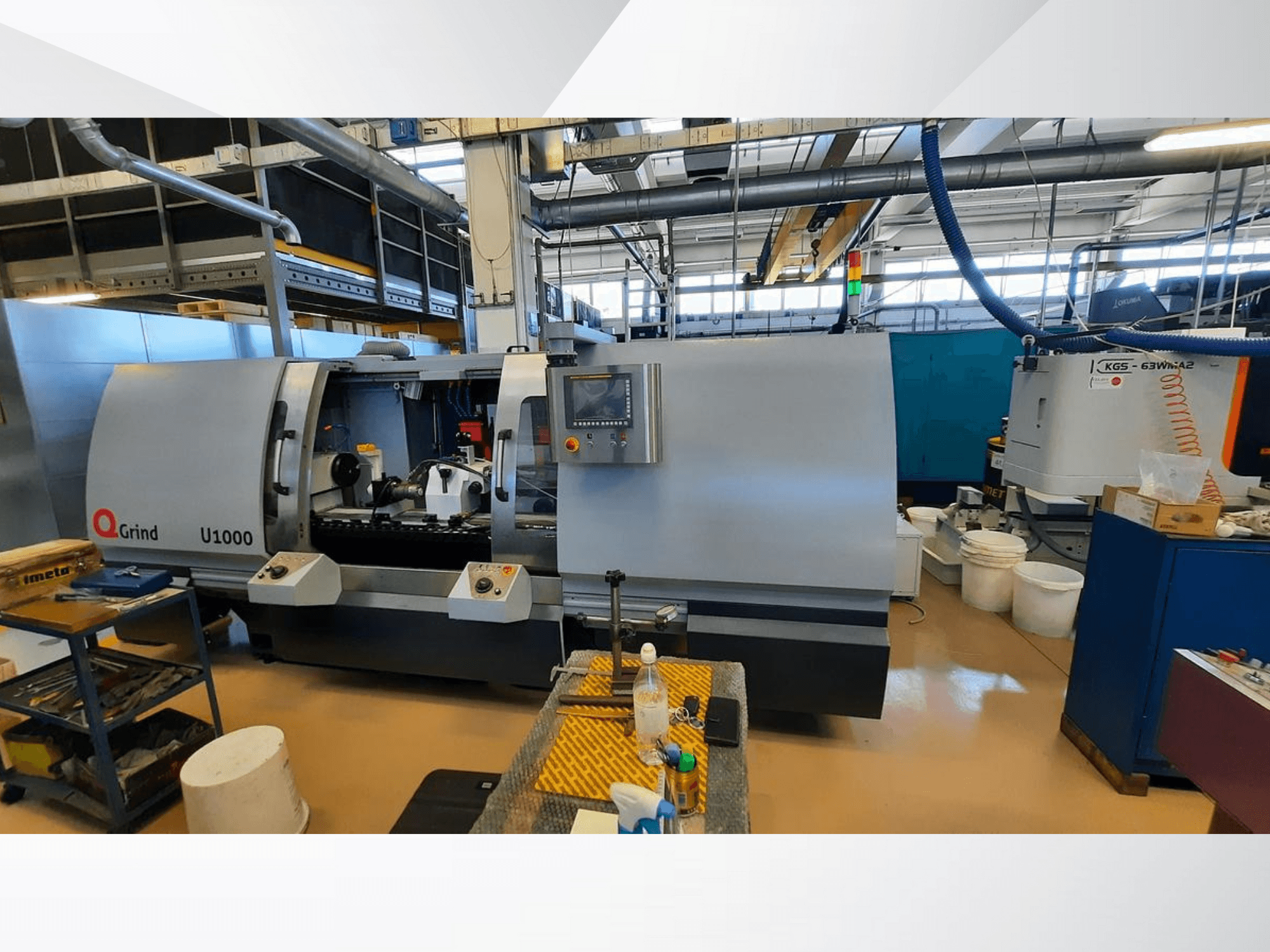 Q-Grind U1000 machine in een werkplaats, vooraanzicht met bedieningspaneel en open snijgedeelte, omringd door apparatuur.