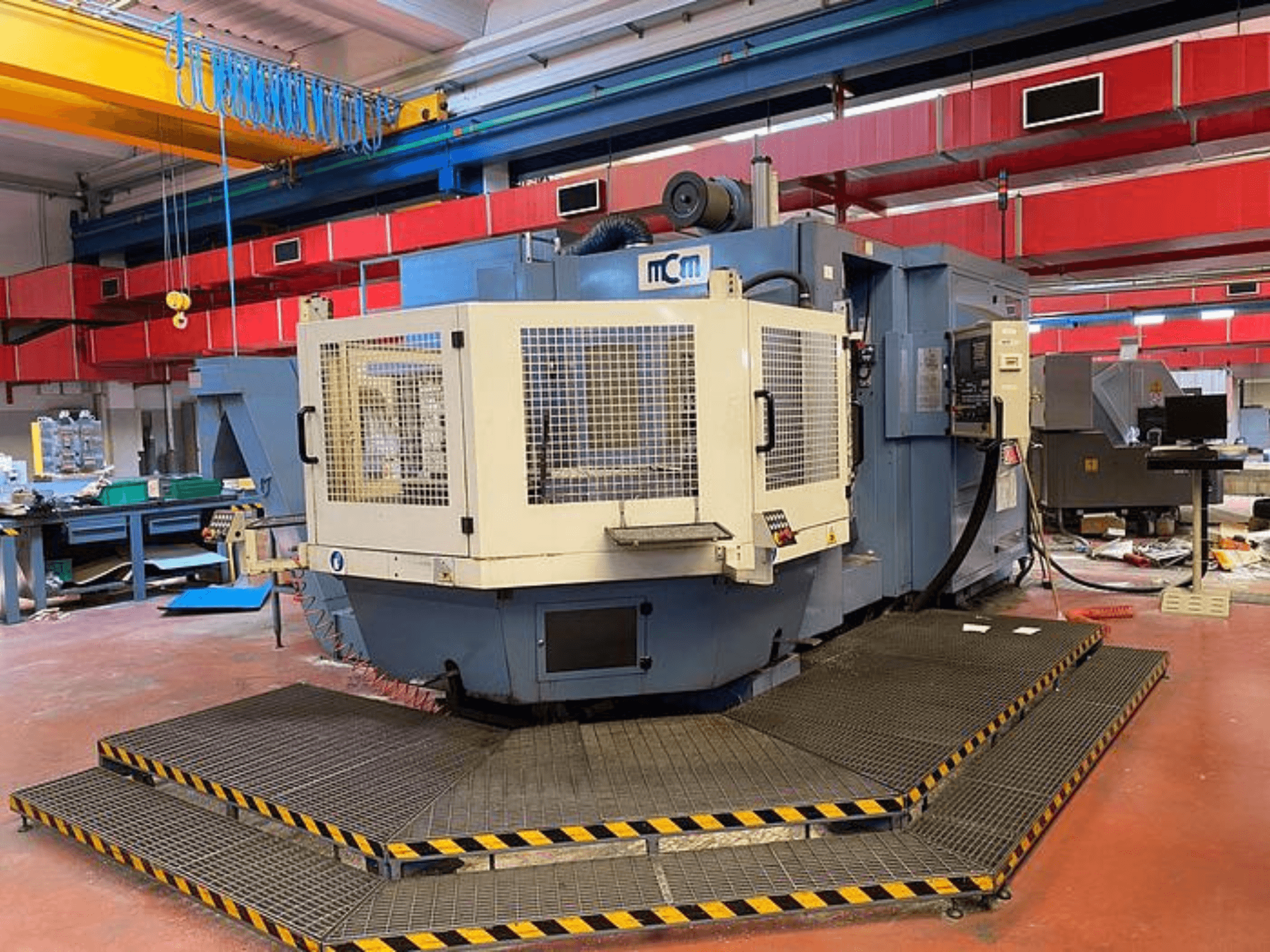 MCM CNC machine in een fabriek, vooraanzicht met beschermkooi, zwarte en gele veiligheidsranden, omringd door industriële apparatuur.