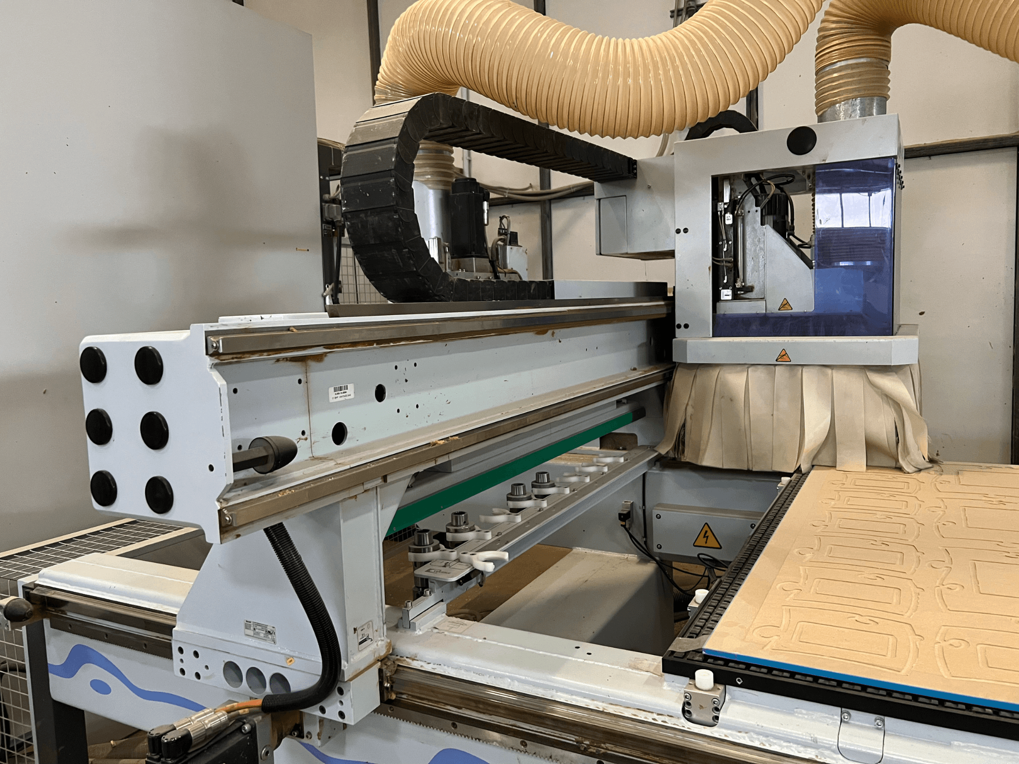 Zijaanzicht van een CNC bovenfrees met een blauw en zilver bedieningspaneel, uitgerust met stofafzuiging en een houtsnijbed.