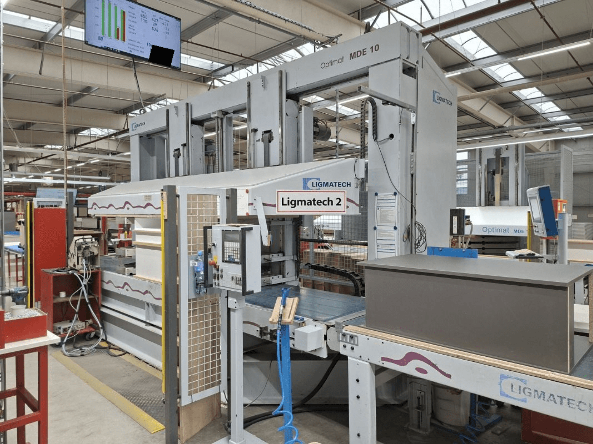 LIGMATECH Optimat MDE 10 machine in een werkplaats, zijaanzicht met bedieningselementen en houtstuk op transportband.