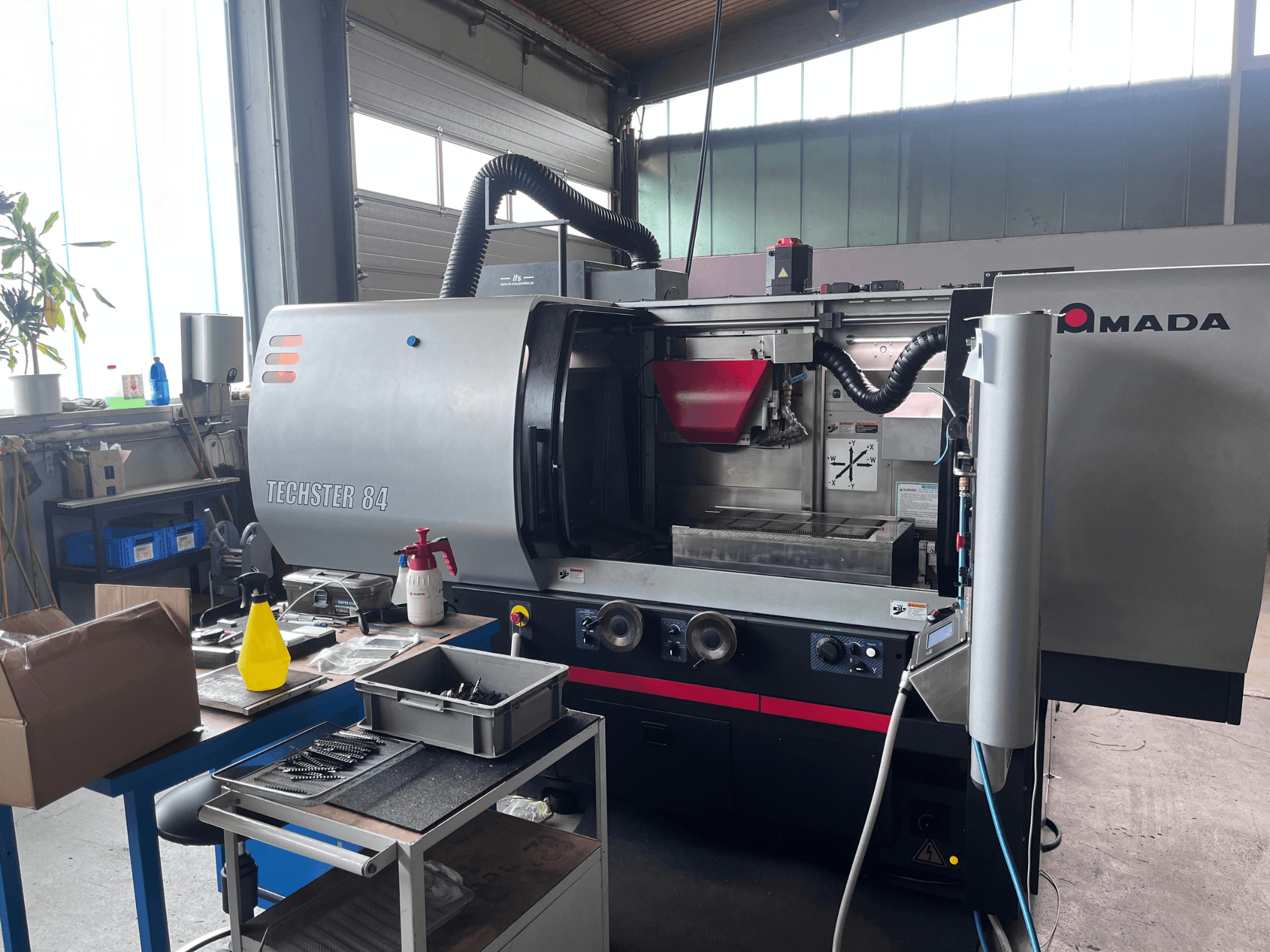 AMADA Techster 84 vlakslijpmachine in een werkplaats, gezien vanaf de voorkant met bedieningselementen en een rode trechter.