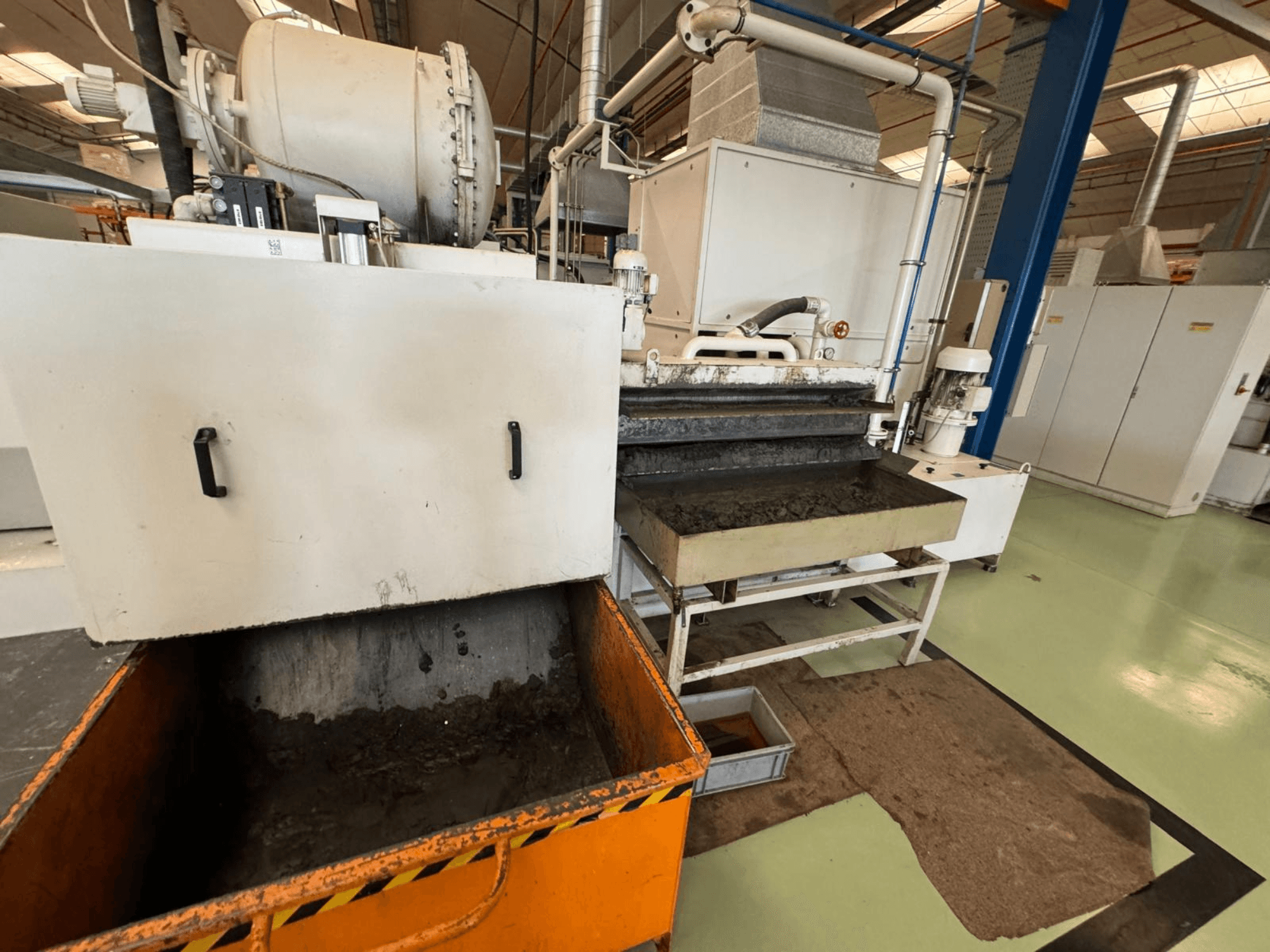 Industriële verwerkingsapparatuur met een oranje afvalbak, vooraanzicht; bevat een motor, transportbanden en onderhoudsruimte.