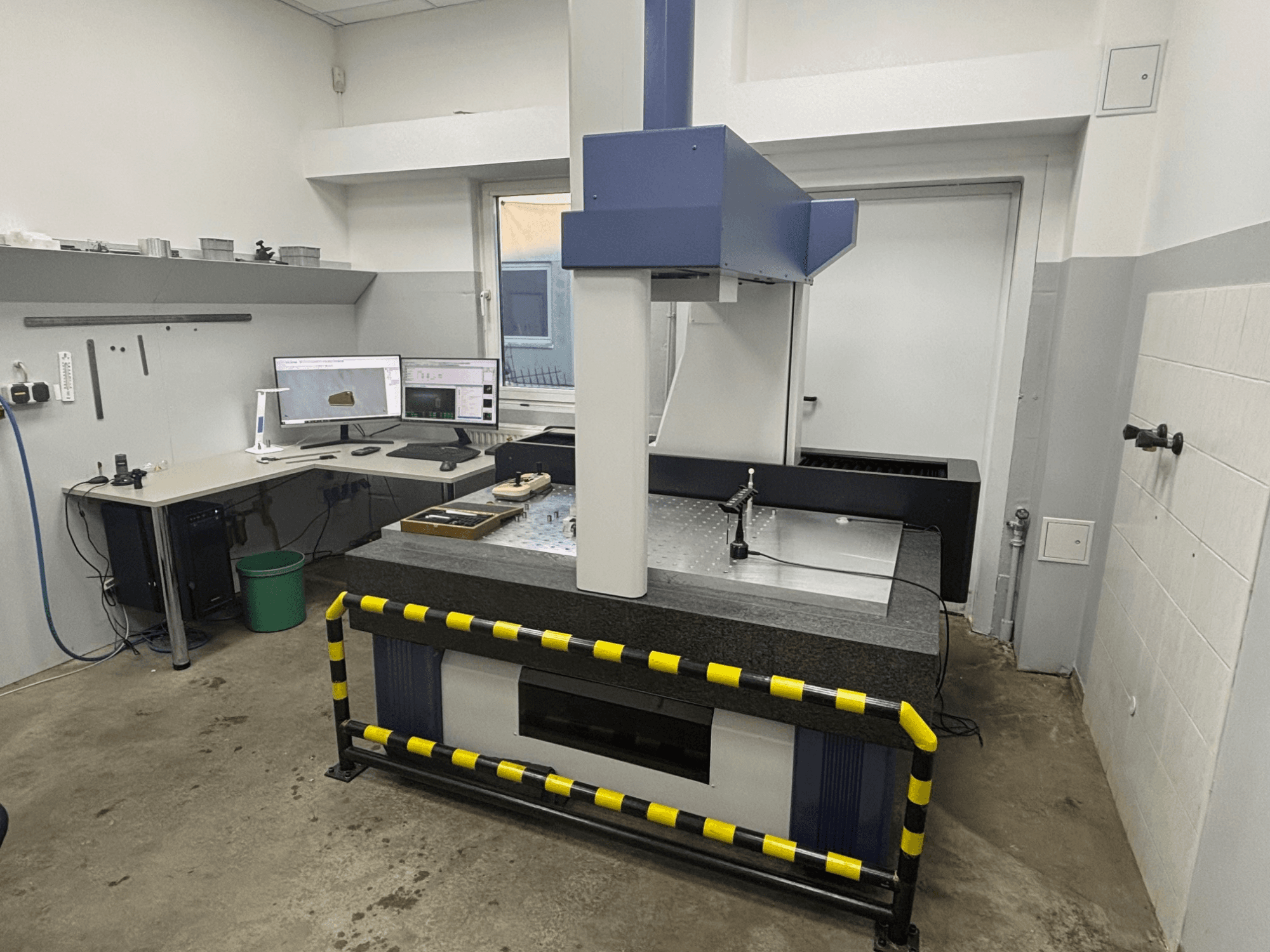 CMM-werkstation met meetmachine, dubbele monitoren en veiligheidshekken. Vooraanzicht met apparatuur en installatiedetails.