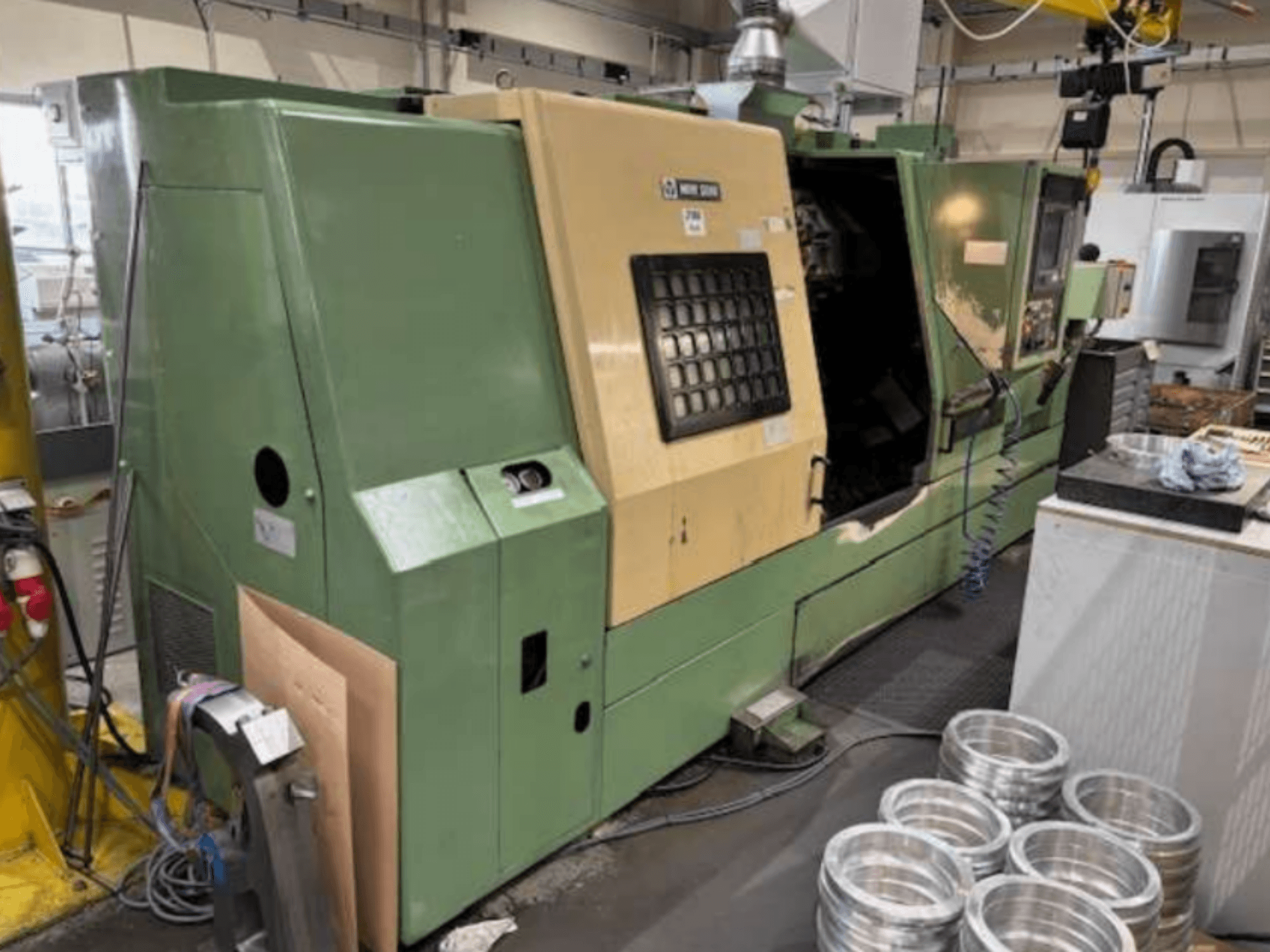 Groene CNC draaibank van Mazak, gezien vanaf de voorkant, met een bedieningspaneel en bewerkte aluminium onderdelen op een werktafel.