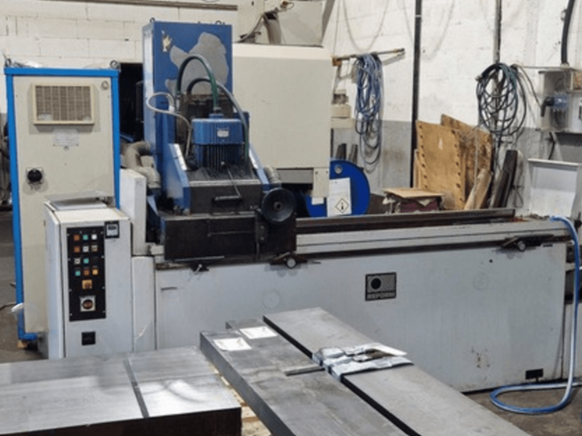 Reform Surface Grinder in vooraanzicht, met het bedieningspaneel, het slijpgebied en de metalen werkstukken op de tafel.