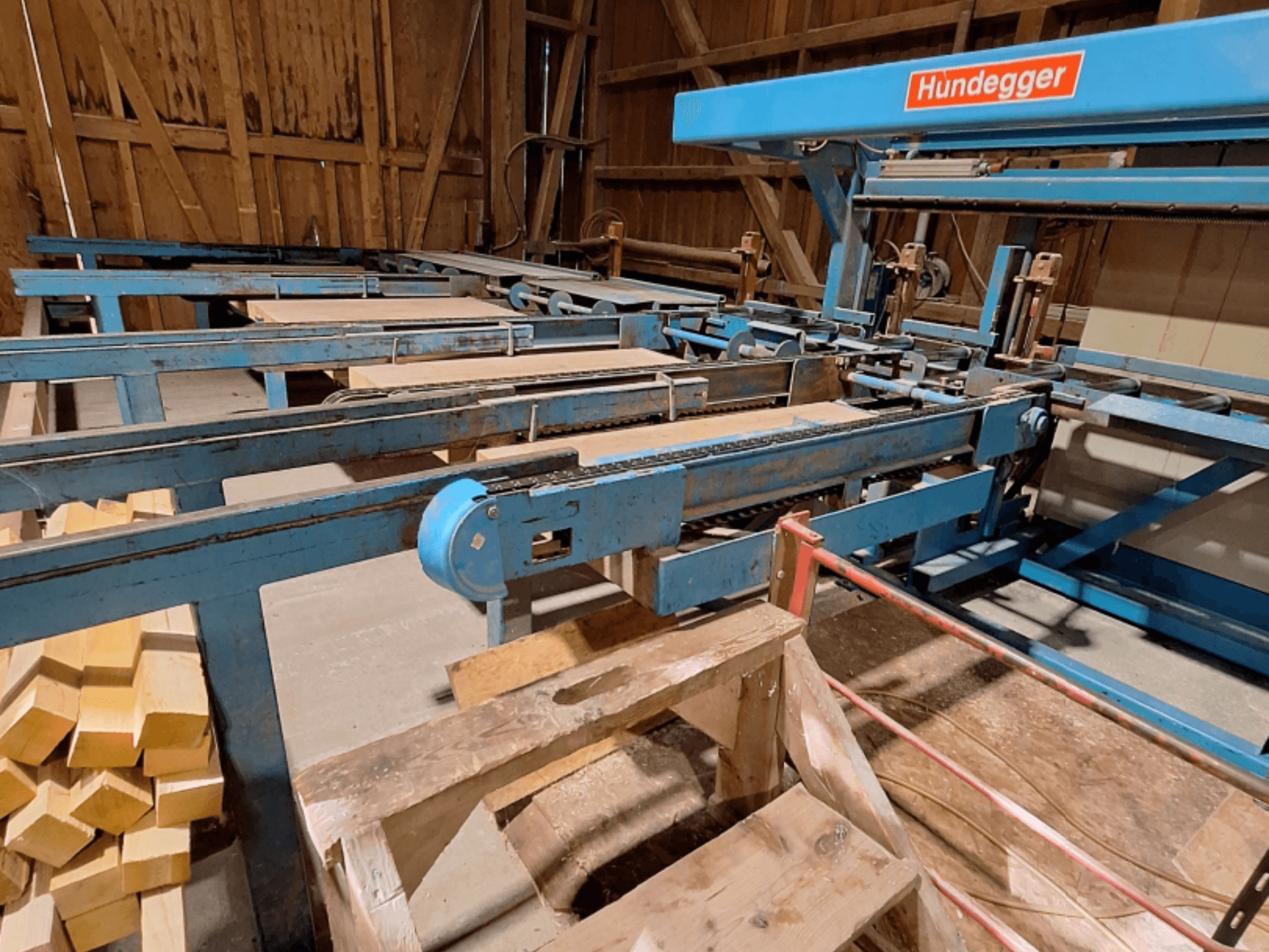 CNC-houtbewerkingscentrum HUNDEGGER K2-A in een schuur, met een blauw metalen frame, een transportband en gestapelde houten balken.
