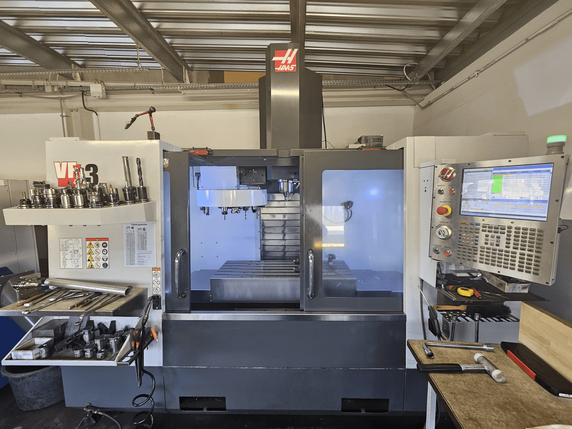 Haas VF-3 CNC machine in een werkplaats, vooraanzicht met gereedschap, bedieningselementen en freesgebied met verlicht interieur.