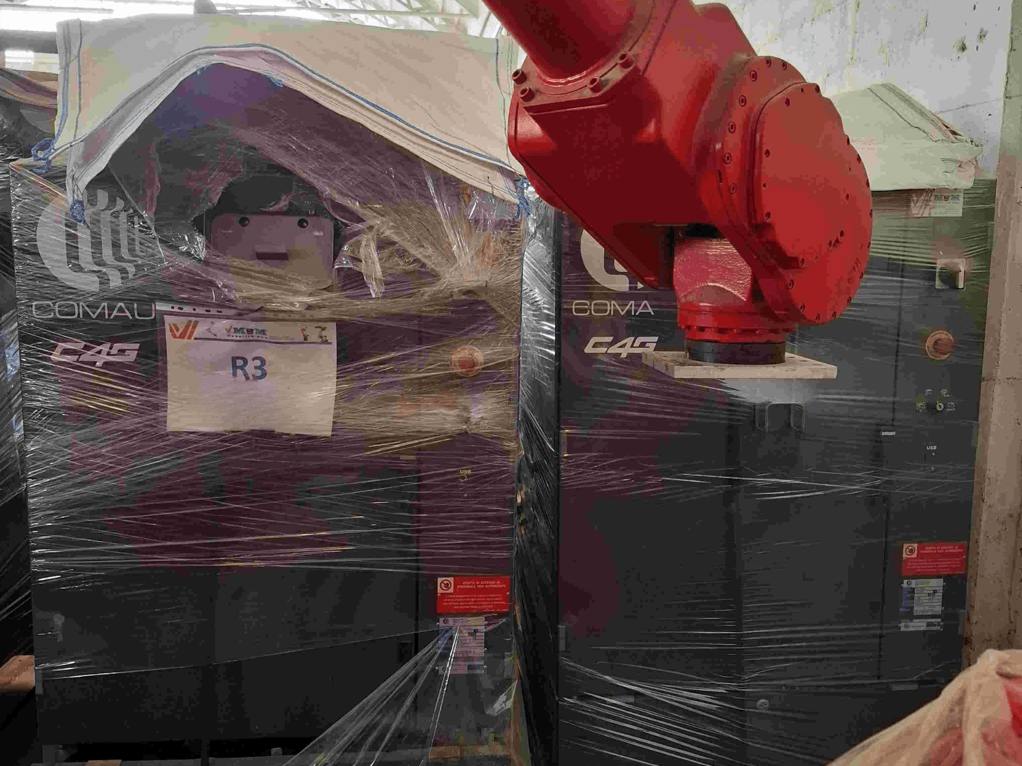 Comau C4G robotarm gezien vanaf de voorkant, verpakt in plastic, met het label "R3" en waarschuwingsstickers zichtbaar op de machine.