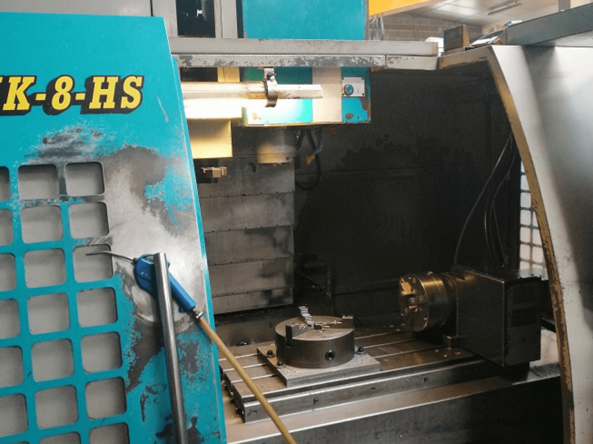 Vooraanzicht van een blauwe SK-8-HS CNC machine, met een werktafel met bankschroef en bedieningspaneel; zichtbare slijtage en vetvlekken.