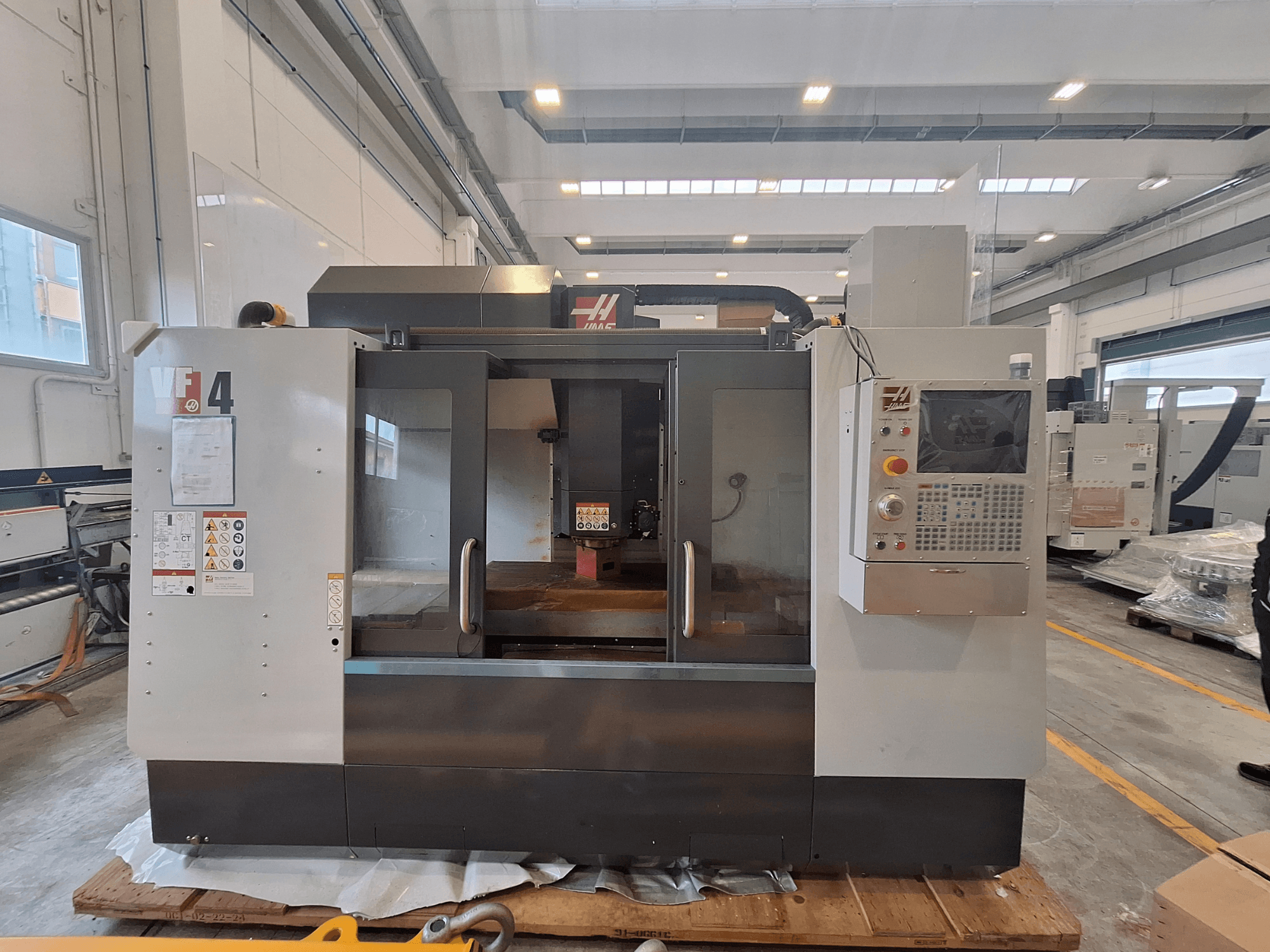 HAAS VF-4 verticaal bewerkingscentrum, vooraanzicht, met het bedieningspaneel, het werkgebied en de veiligheidslabels in een magazijnomgeving.