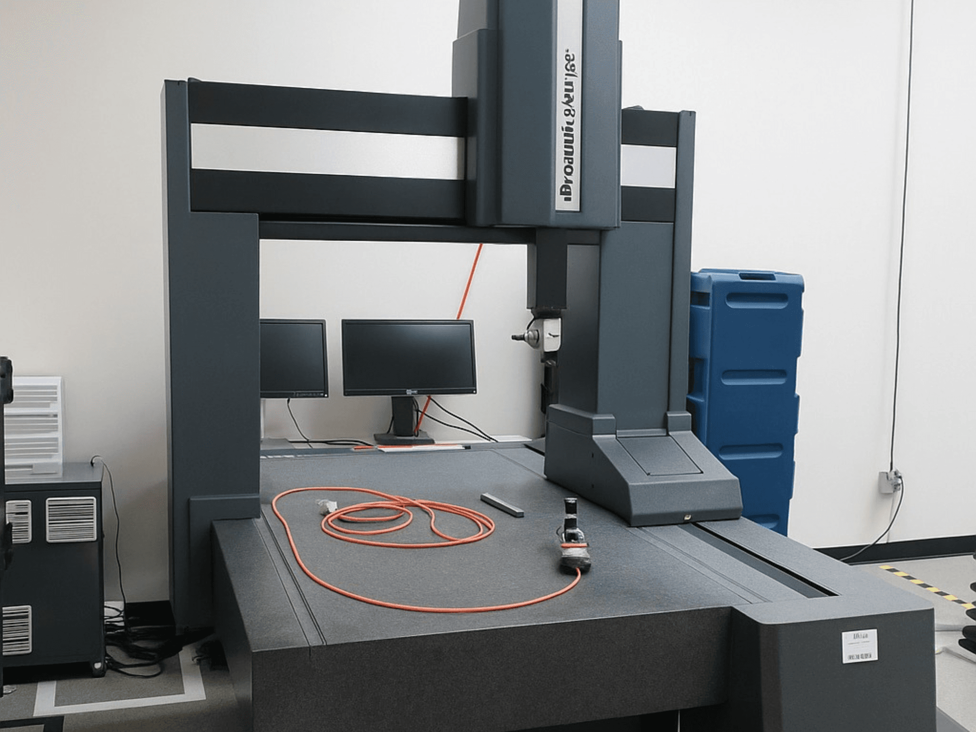De coördinaten meetmachine van Brooks Automation met twee monitoren, vooraanzicht, heeft een grijs tafelblad en meetgereedschap.