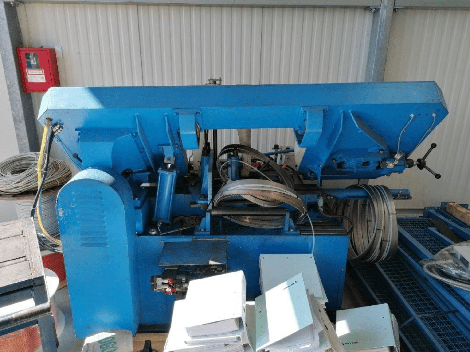 Blauwe industriële machine met hydraulische onderdelen, zijaanzicht, met slangen en platen metaal op een oppervlak.