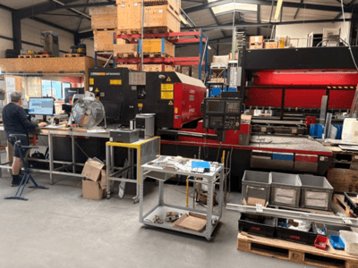 Een AMADA LC-2415αIII CO₂-lasersnijmachine in een werkplaatsomgeving, met op de voorgrond een computerwerkplek.