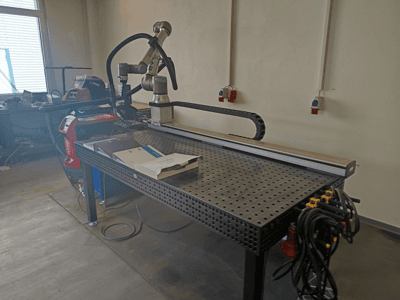 FRONIUS TransSteel 4000-pulslascel, gezien vanaf de zijkant, met een robotarm, werkruimte en stroomaansluitingen.