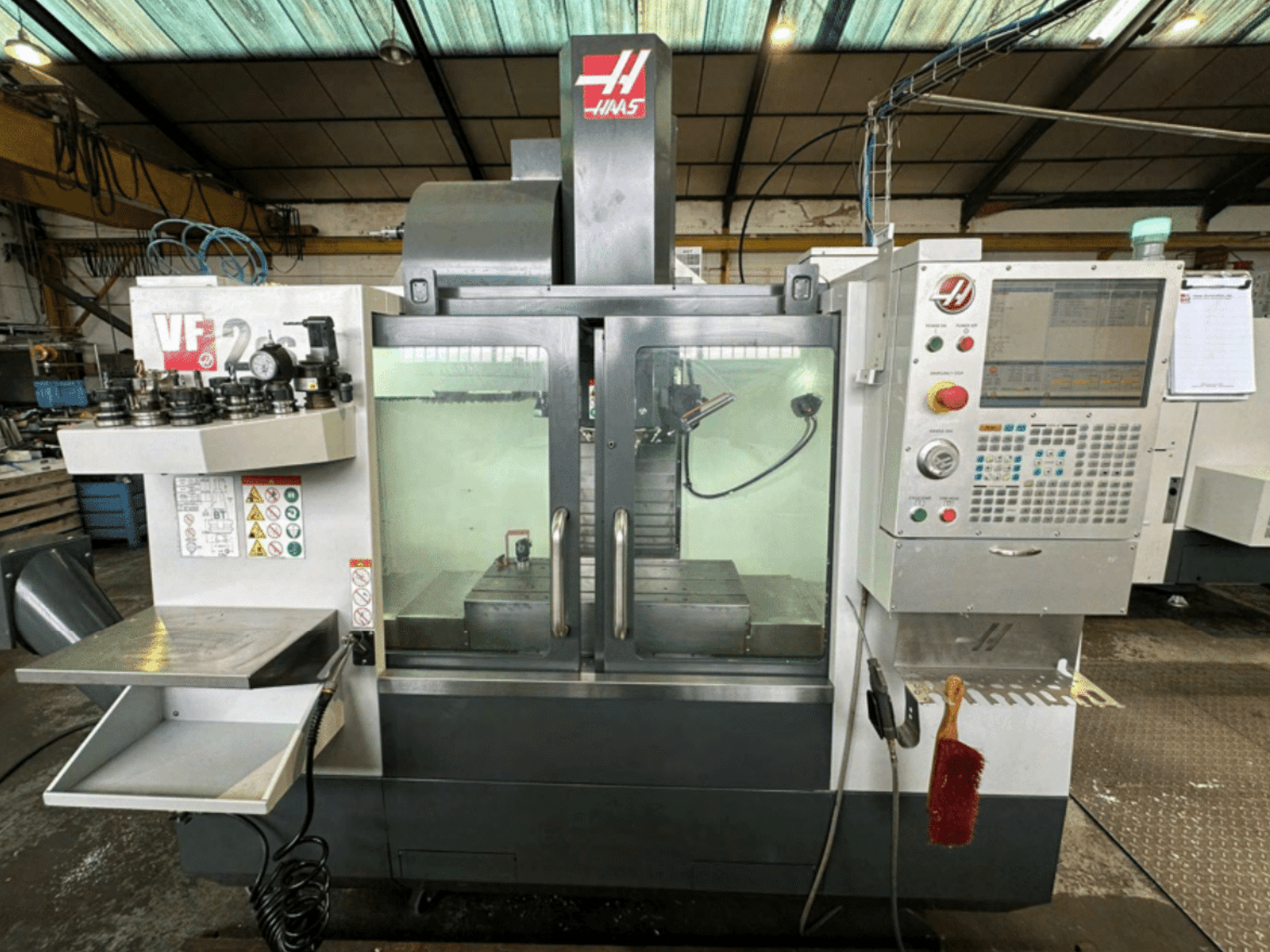 Vooraanzicht Haas VF2 CNC machine, met bedieningspaneel, gereedschaphouders en werkruimte met veiligheidslabels en een borstel.