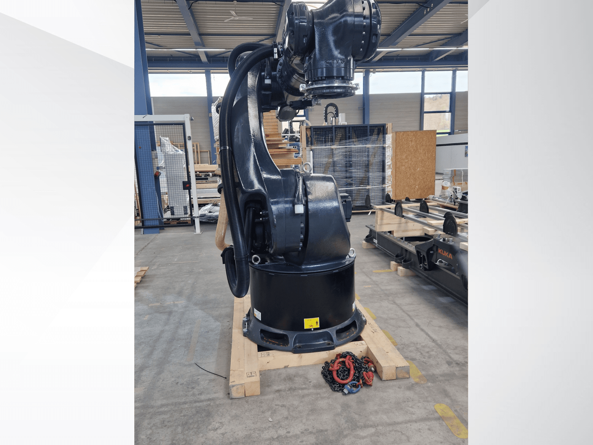 KUKA KR 340 R3330-robotarm in een magazijn, zijaanzicht waarin de voet en de kabels te zien zijn, met kettingen op de voorgrond.