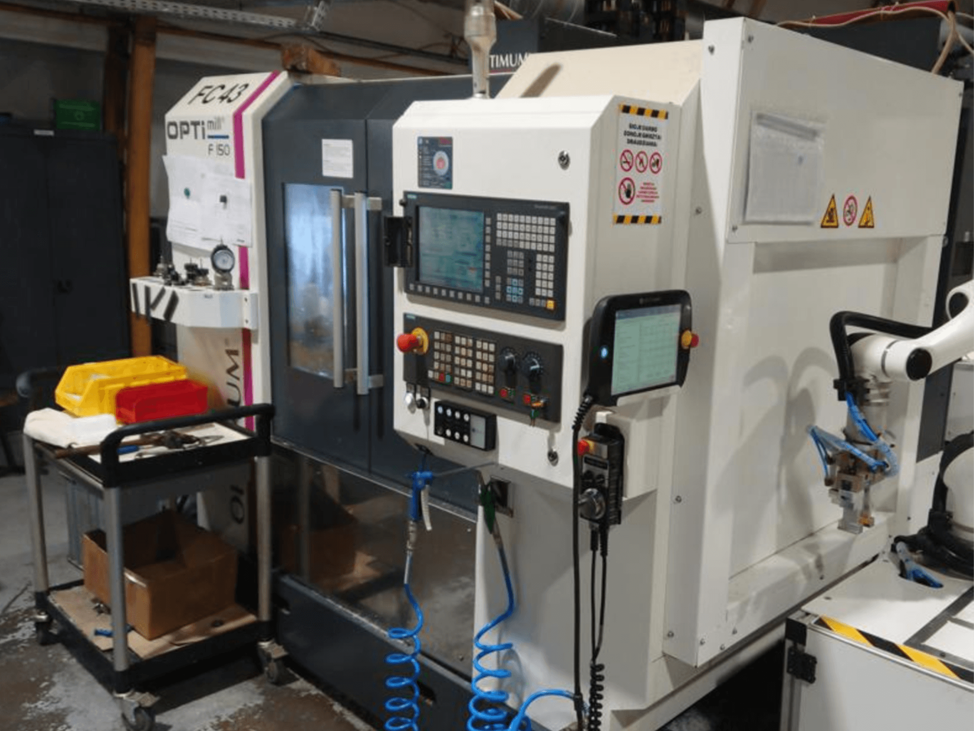 Bedieningspaneel van OPTIMUM F50 freesmachine met slangen, gereedschappen en een robotarm zichtbaar in een werkplaats.