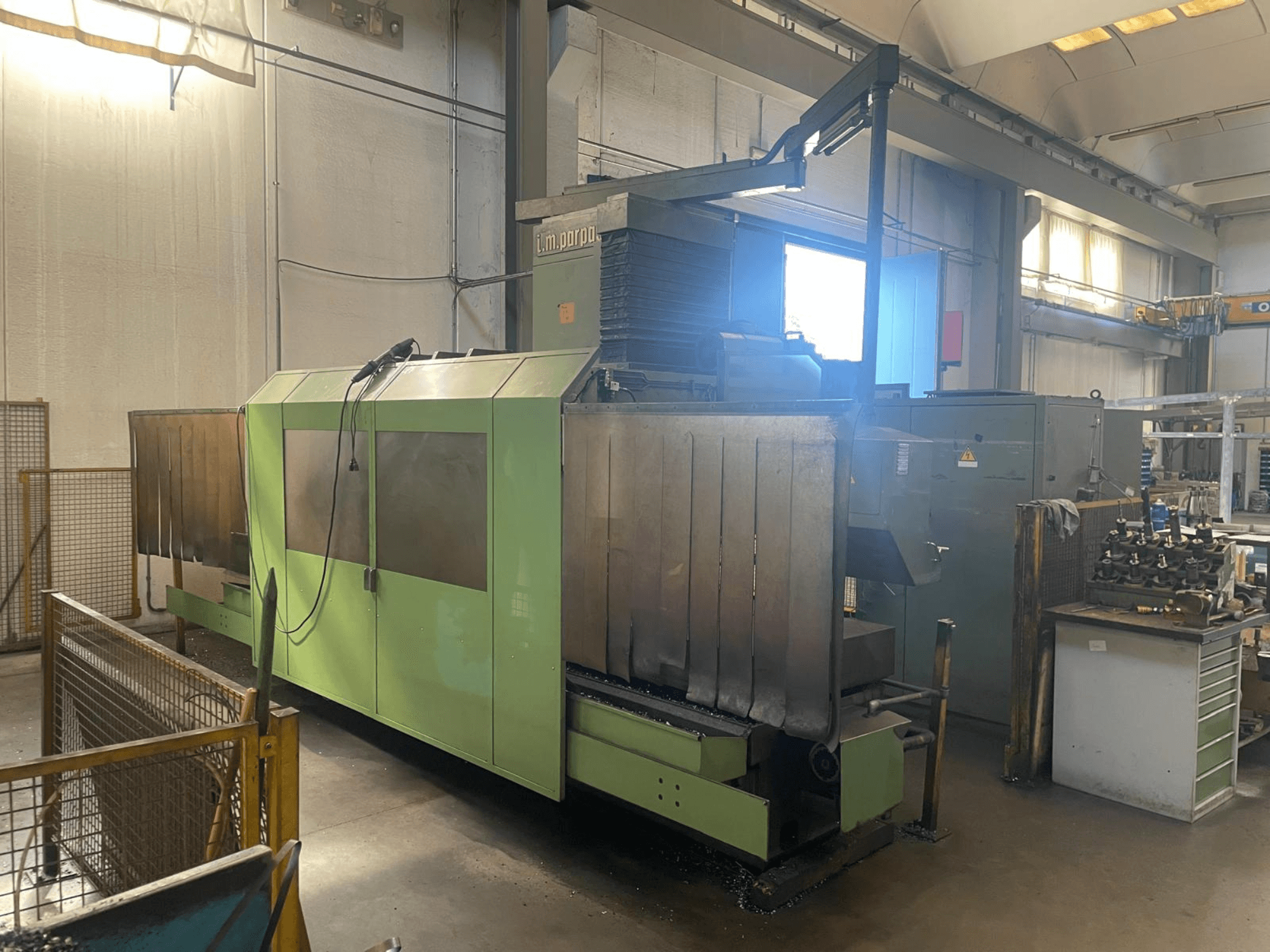 Groene industriële machine met zichtbare metalen onderdelen, vooraanzicht; voorzien van bedieningspaneel, veiligheidswaarschuwingen en werkstation in de buurt.