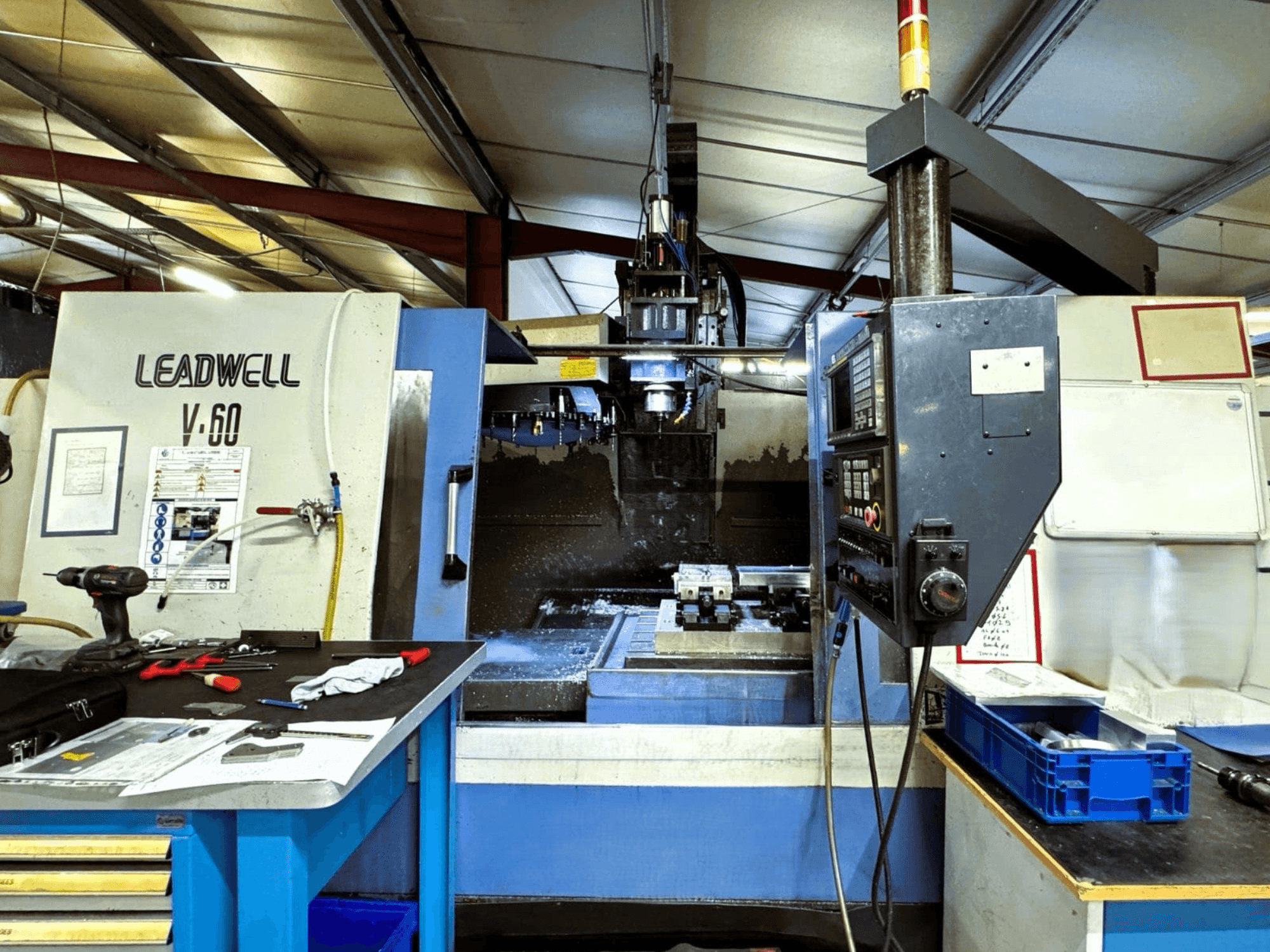 Het verticale bewerkingscentrum Leadwell V-60 gezien vanaf de zijkant, met het bedieningspaneel en het werkgebied met gereedschappen en onderdelen.