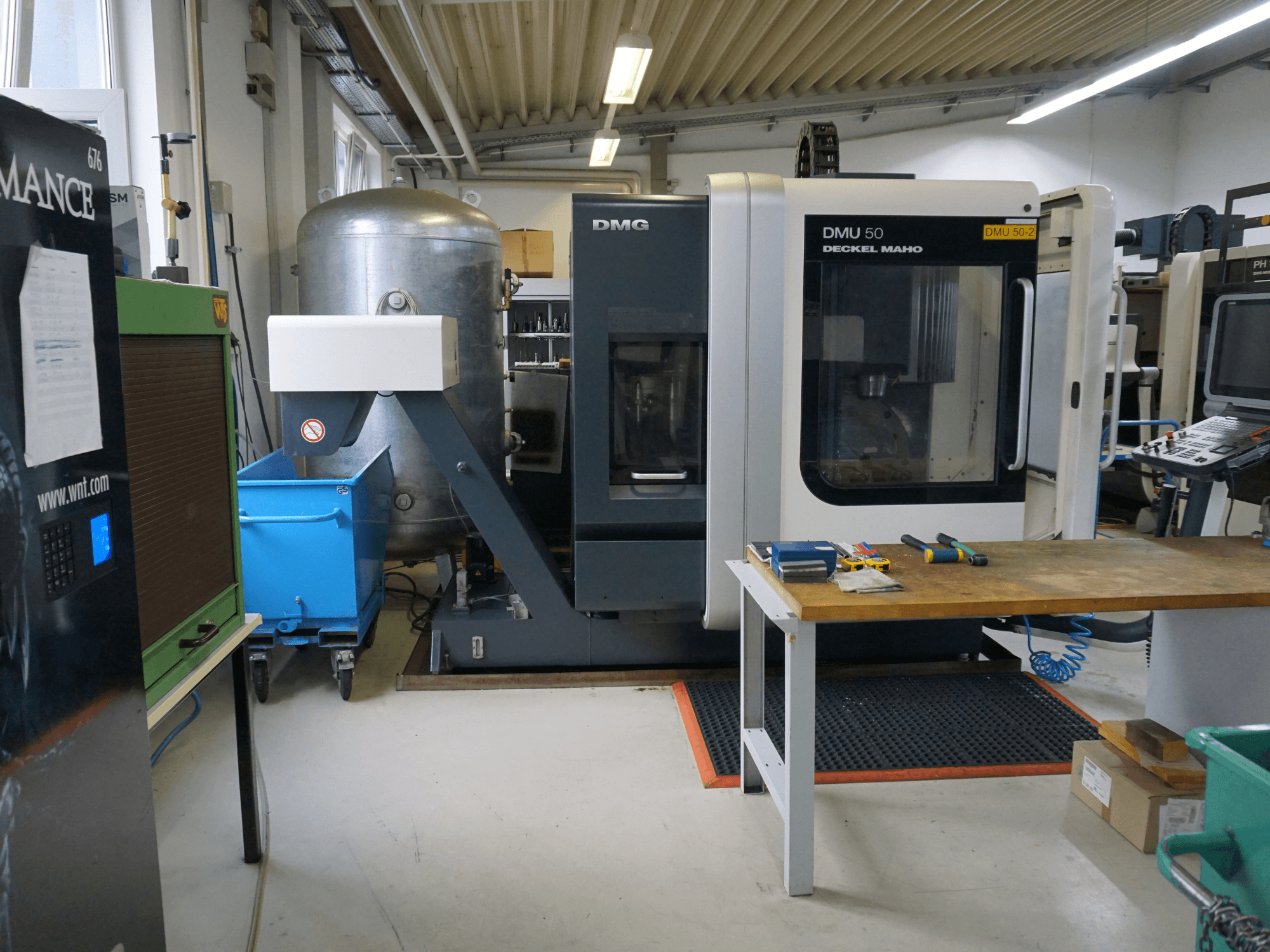 DMG DMU 50 freesmachine in een werkplaats, vooraanzicht, met gereedschap op een houten tafel en een blauwe bak op wielen vlakbij.