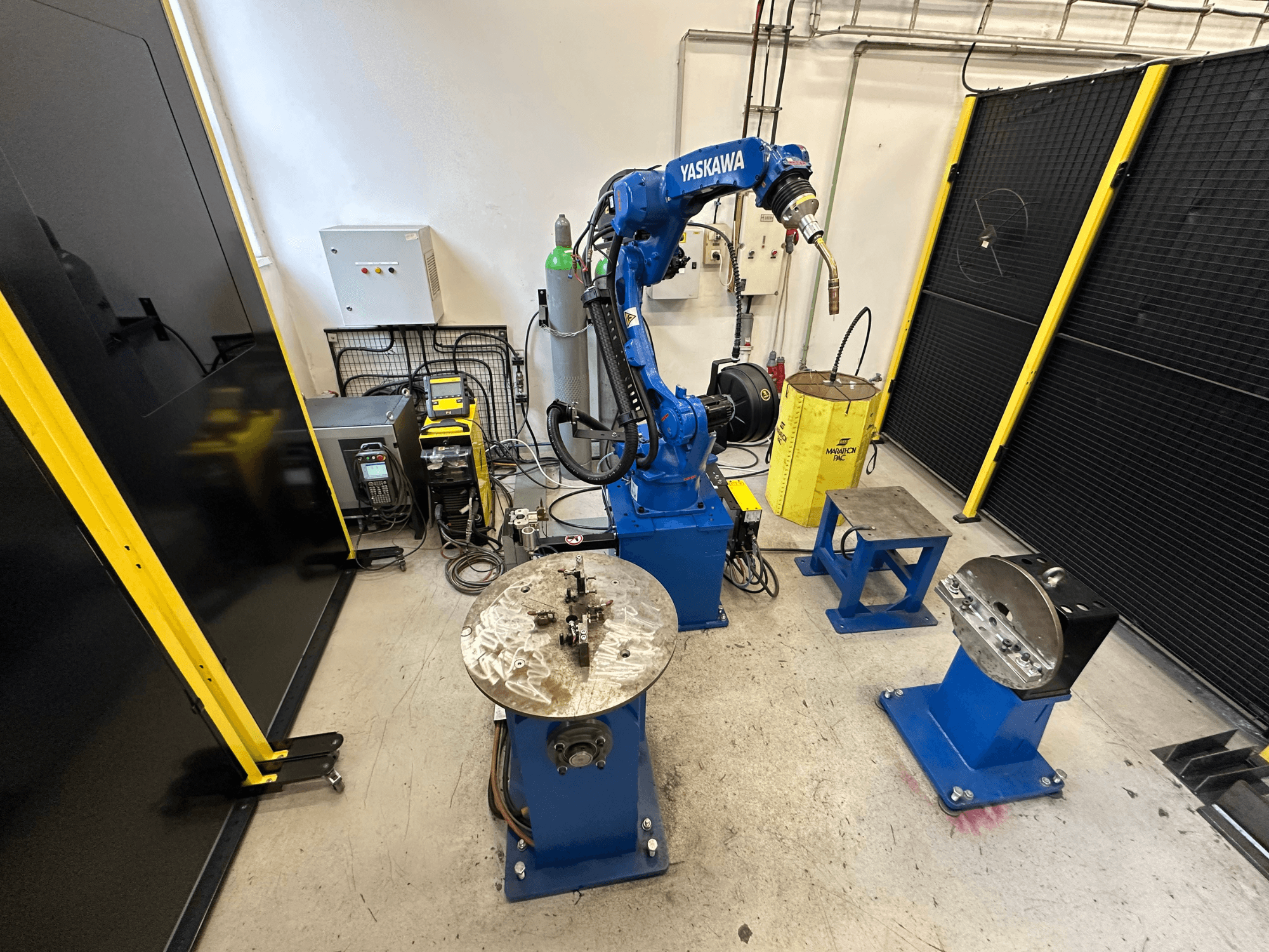 Yaskawa robotarm in blauw met twee werktafels, lasapparatuur en veiligheidshekken; vooraanzicht industriële werkruimte.