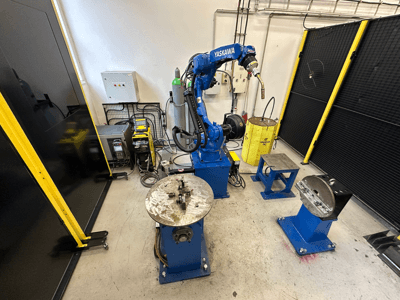 Yaskawa robotarm in blauw met twee werktafels, lasapparatuur en veiligheidshekken; vooraanzicht industriële werkruimte.