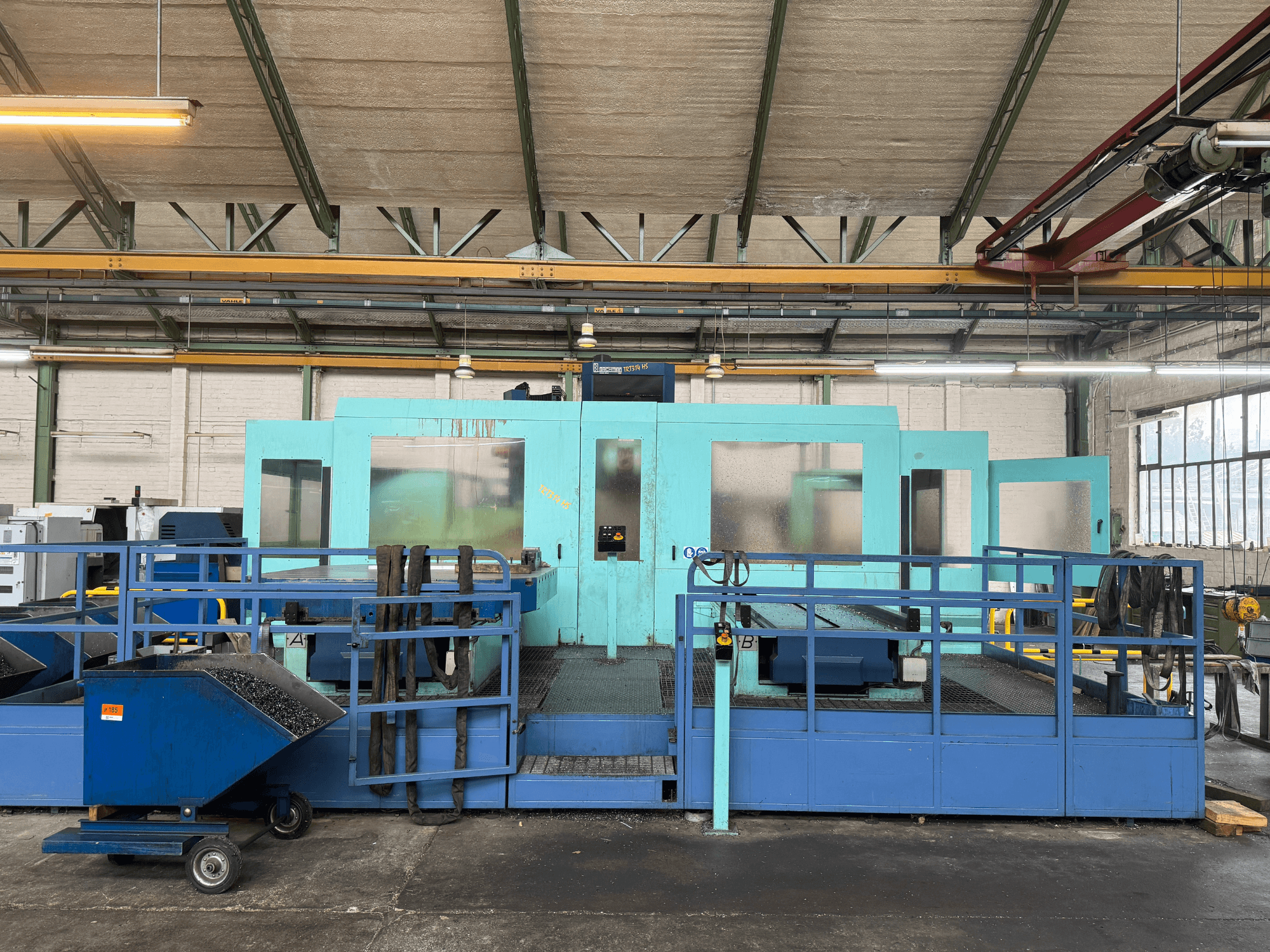 Industriële blauwe CNC machine met beschermpanelen, vooraanzicht, met opbergruimte voor gereedschap en een afvalverzamelkar aan de zijkant.