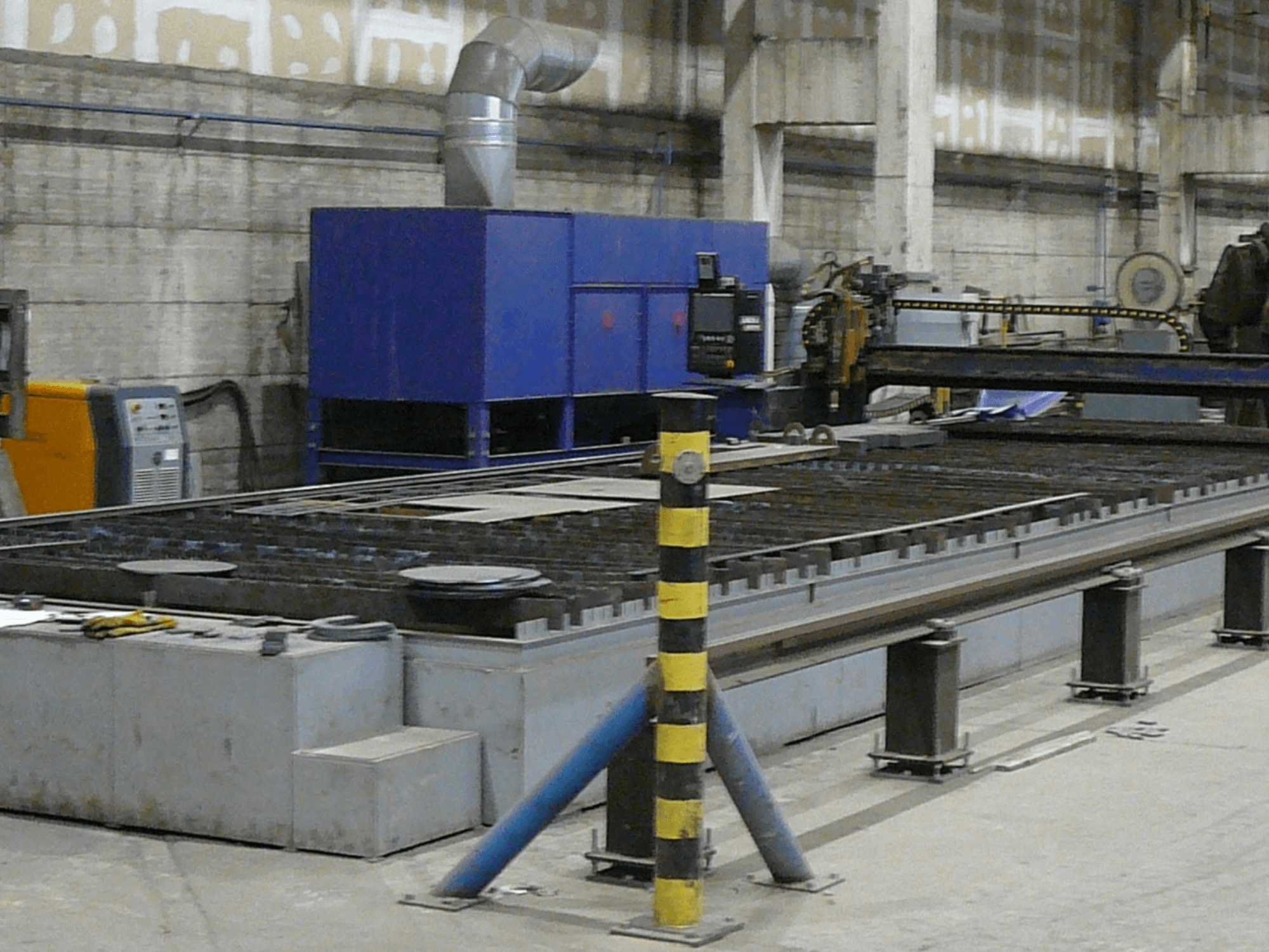 Opstelling industriële metaalsnijmachine, zijaanzicht, met een blauw bedieningskastje, veiligheidsstrepen en een metalen werkblad.