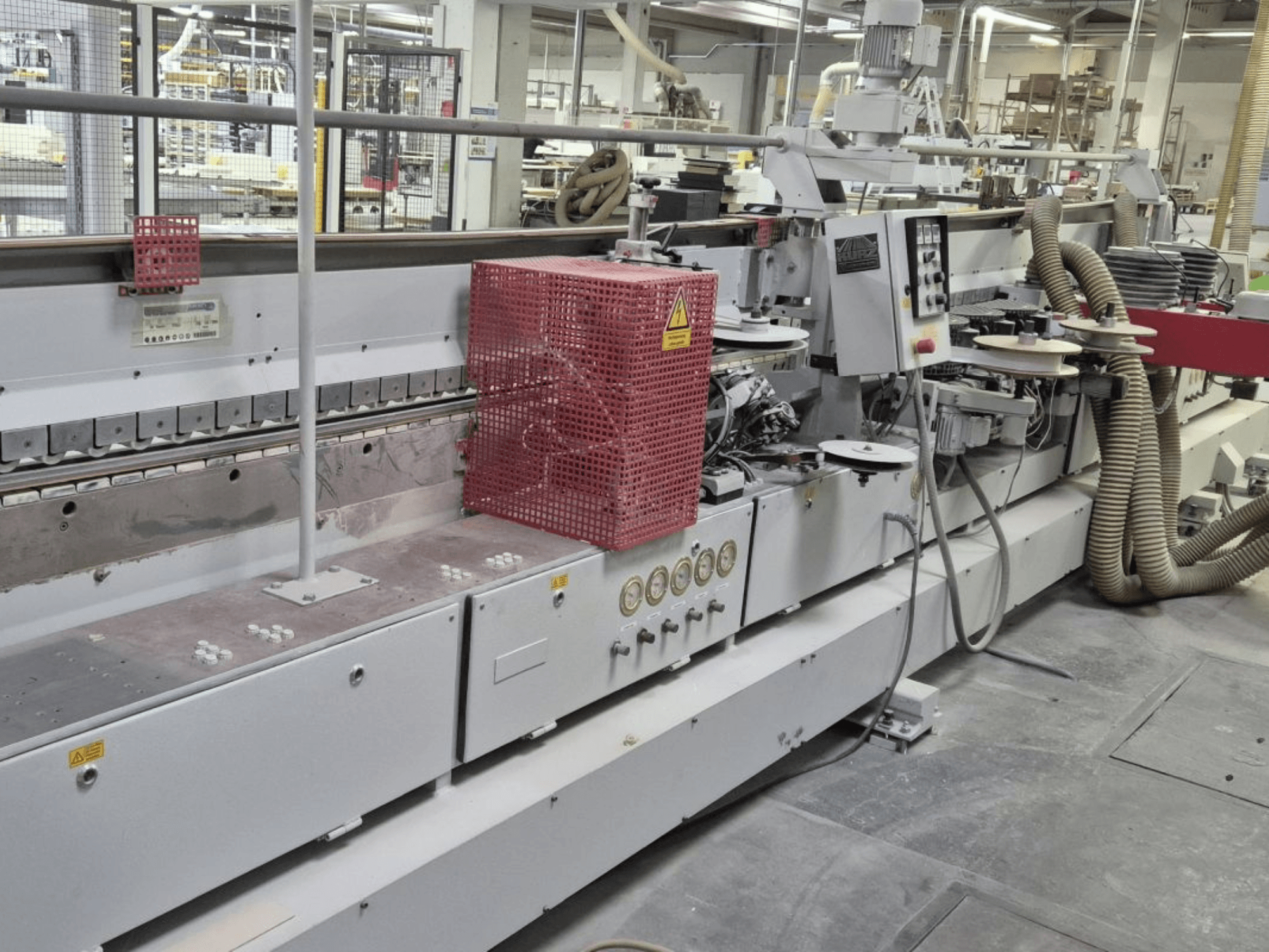 Kantenaanlijmmachine met bedieningspaneel, zuigslangen en een rode veiligheidskap, gezien vanaf de zijkant.