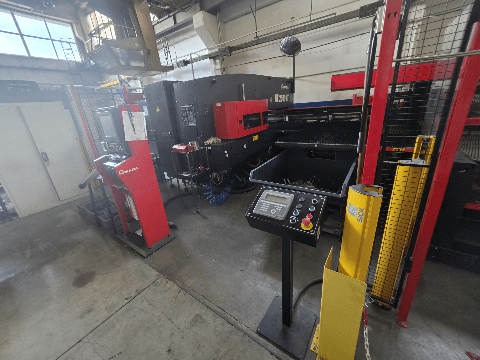 CNC-ponsmachine AMADA AE-2510NT, vooraanzicht met bedieningspaneel, veiligheidsvoorzieningen en de omringende industriële omgeving.