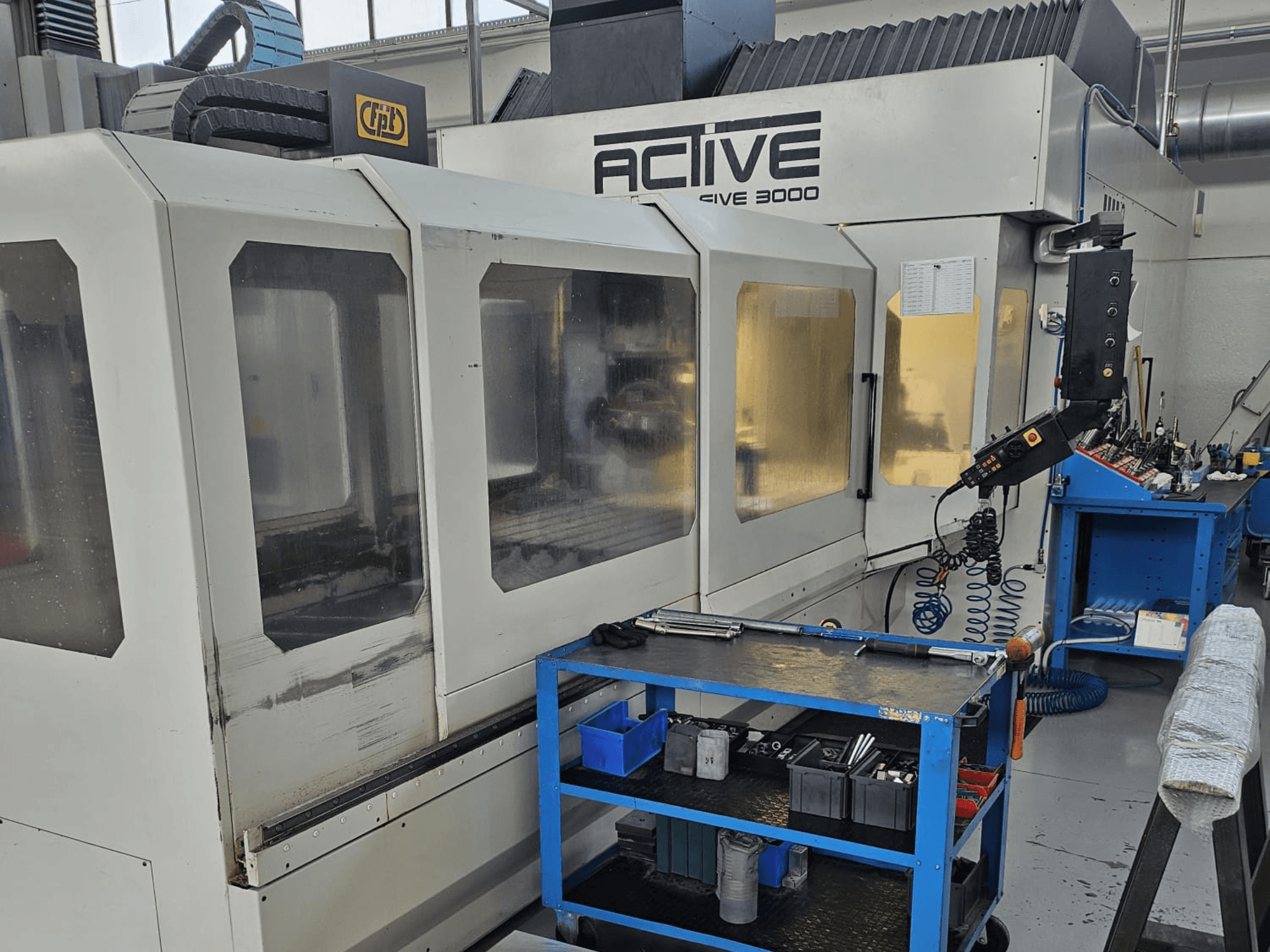 Actieve 3000 CNC machine in een werkplaats, vooraanzicht met gereedschappen op een blauwe kar, zichtbare bedieningselementen en werkgebied binnenin.
