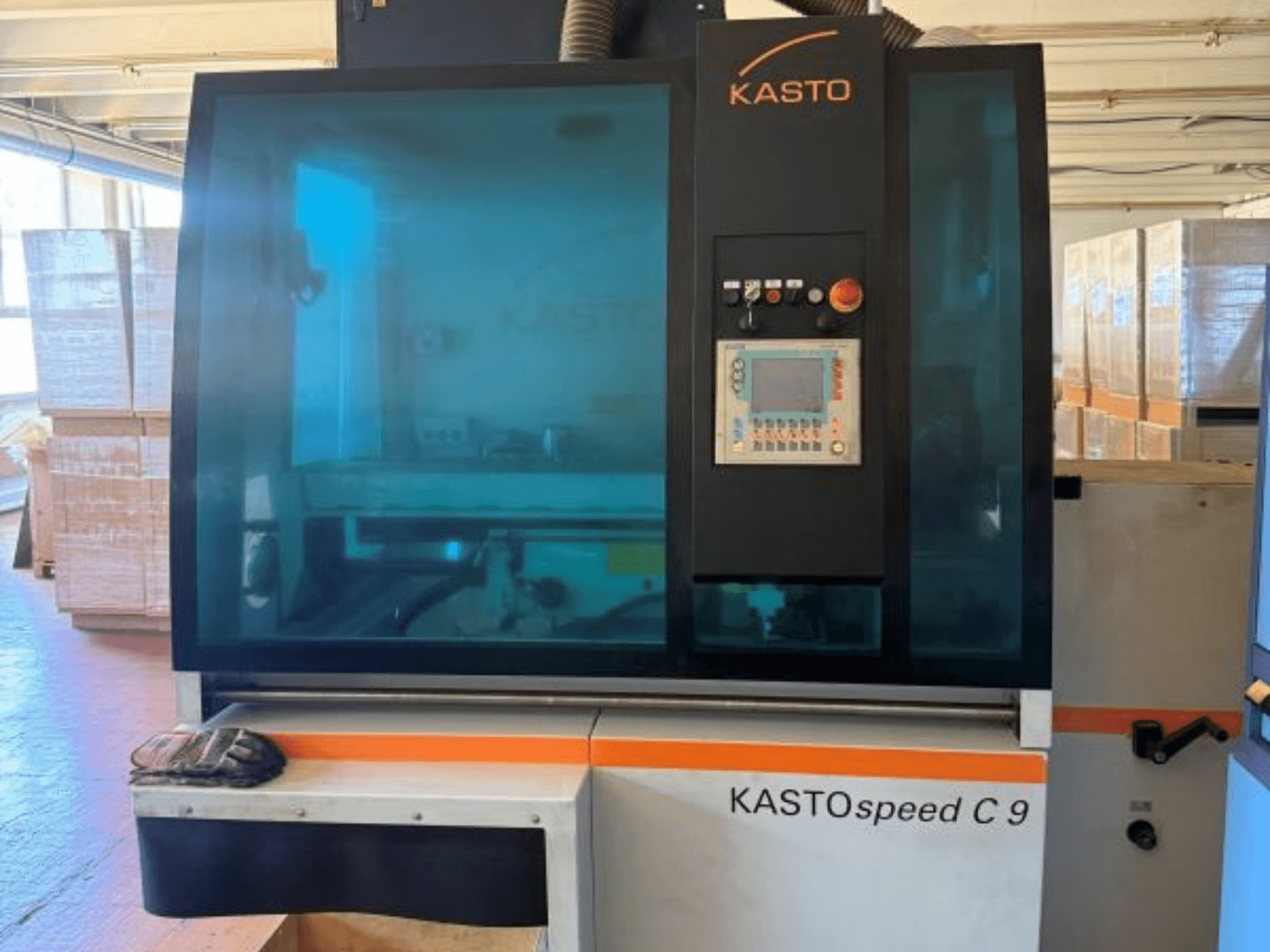 Vooraanzicht van de KASTO KASTOspeed C 9 cirkelzaag voor metaal, met bedieningspaneel en veiligheidsglas, met houten kratten op de achtergrond.