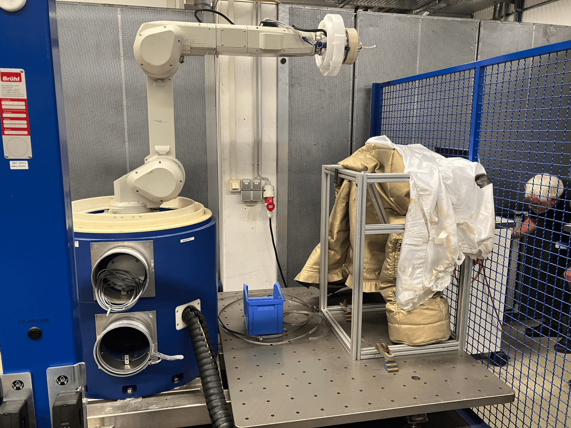 Robotarm bij een blauwe kamer, met een wit en goudkleurig pak op een metalen frame, zijaanzicht met een achtergrond van veiligheidsgaas.