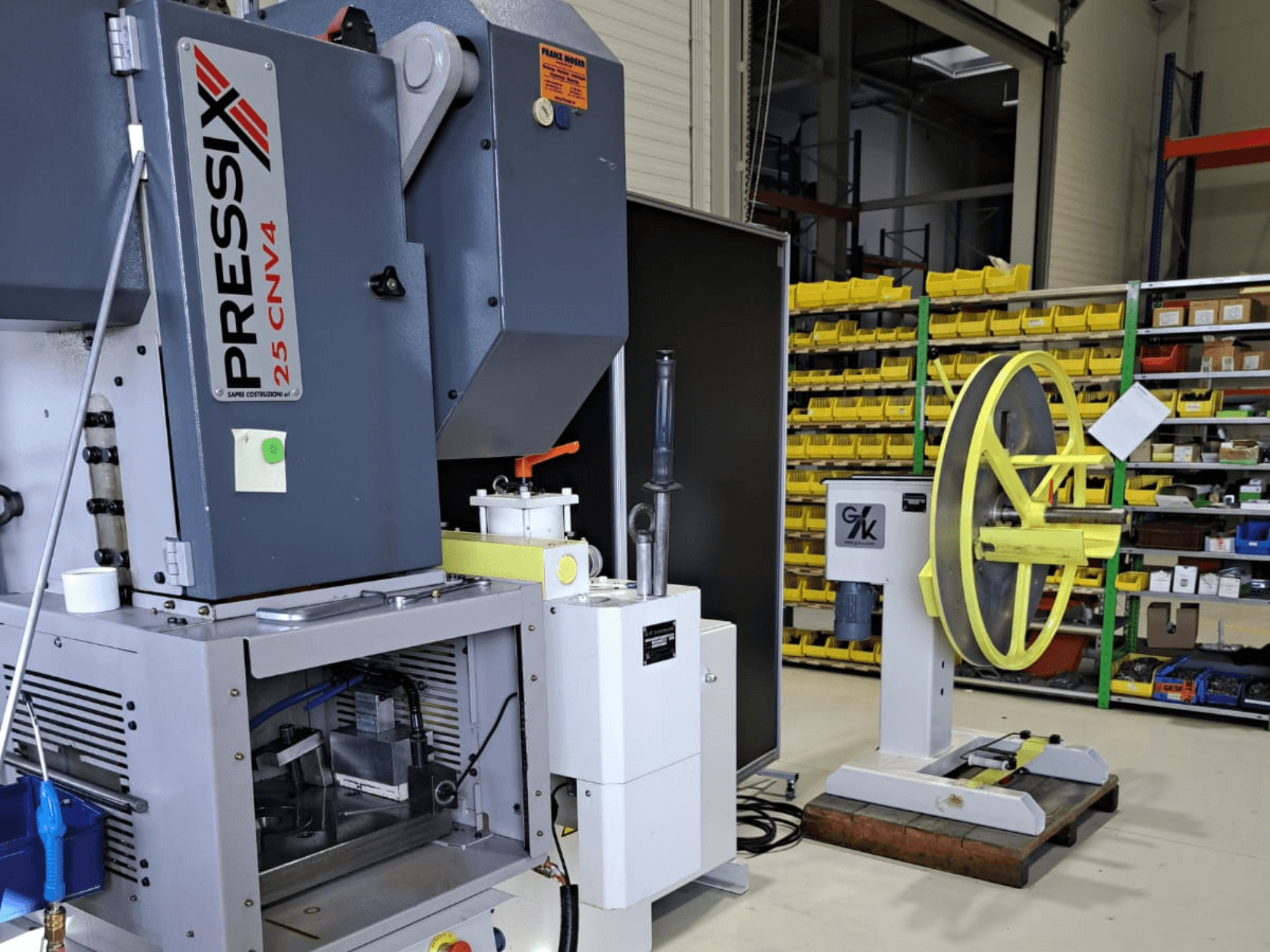 Industriële machineopstelling met links de PRESSIX 25 CNV4 en rechts een geel wiel gemonteerd op een grijze voet, vooraanzicht.