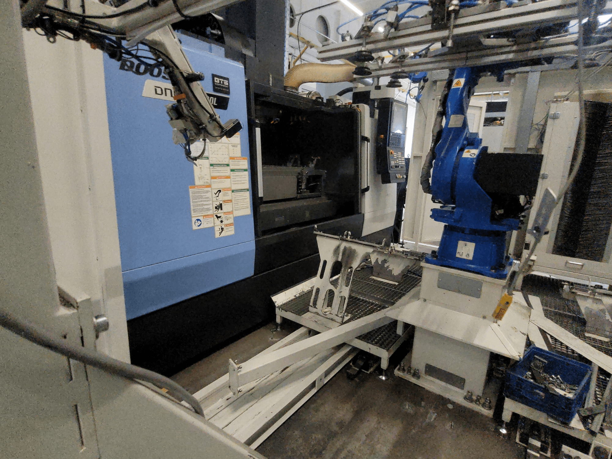 DOOSAN DNM 6700L verticaal bewerkingscentrum in zijaanzicht, met zichtbare robotarm en bedieningspaneel, waarmee de opstelling van de machine wordt getoond.