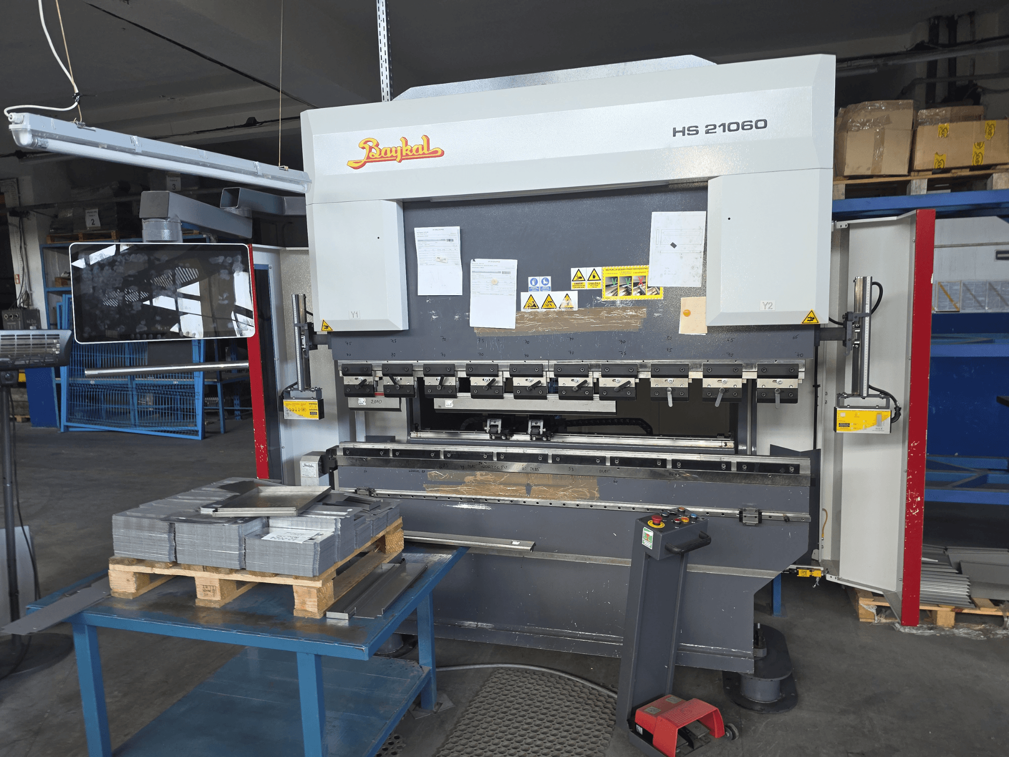 Baykal APHS 21060 kantpers in een werkplaats, zijaanzicht met bedieningspaneel, gestapelde metalen platen en veiligheidsborden.