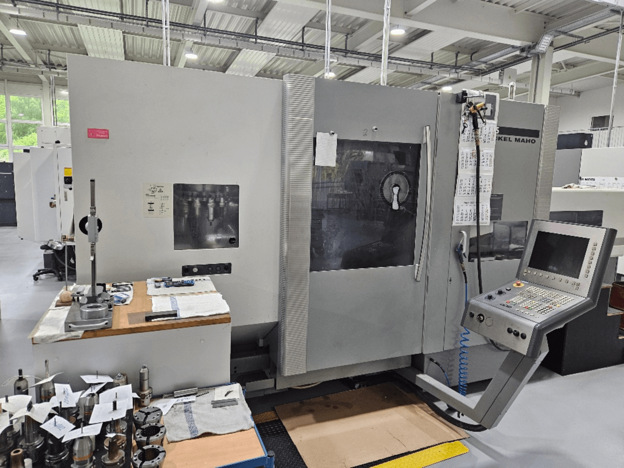 Een vooraanzicht van een DMG Mori DMU 70 machine met bedieningspaneel, gereedschappen op een houten tafel en verschillende werkstukken in de buurt.