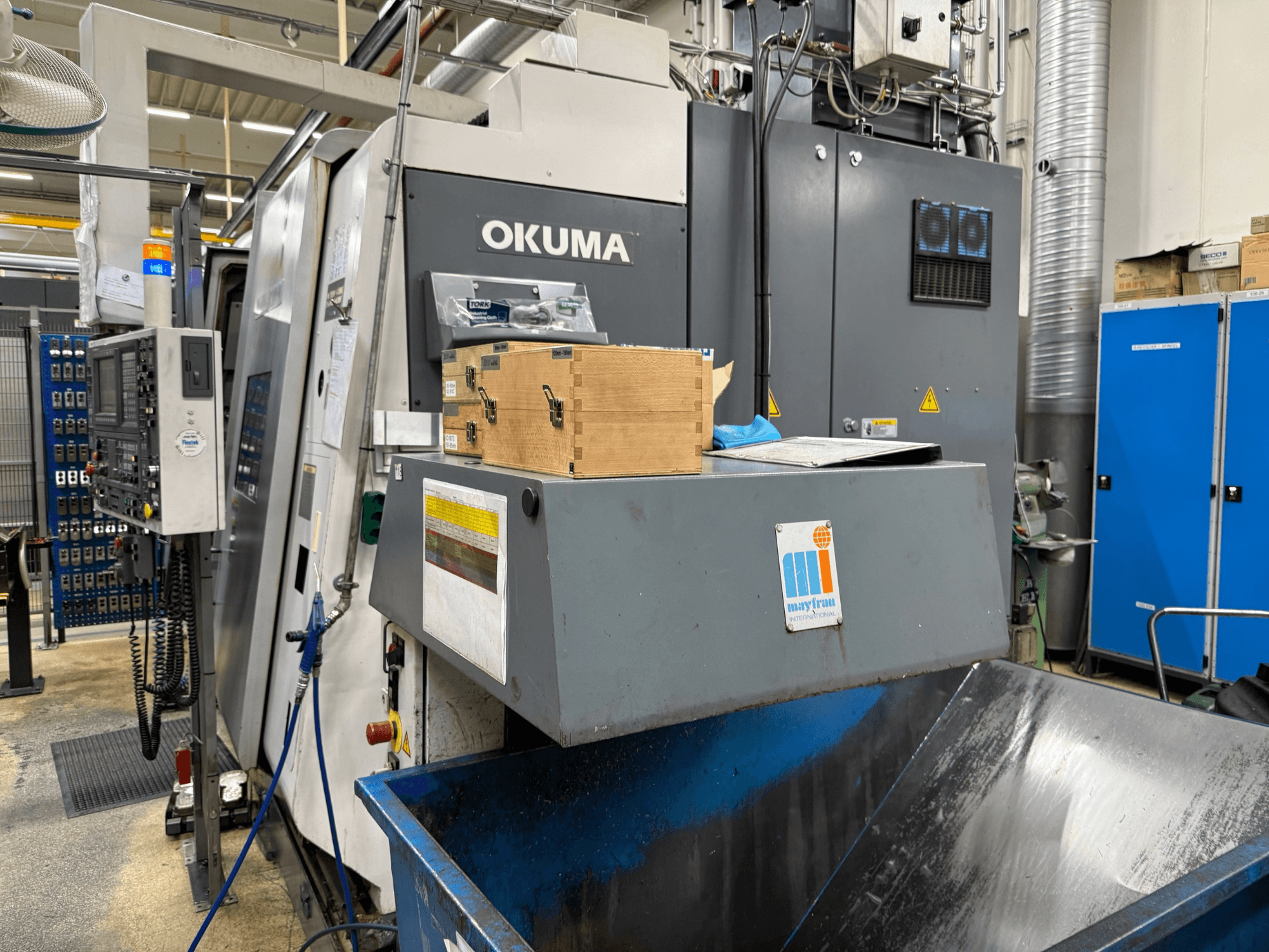 Okuma CNC machine in de fabriek, vooraanzicht; met bedieningspaneel, houten kisten en blauwe afvalbak met Mayfran logo.