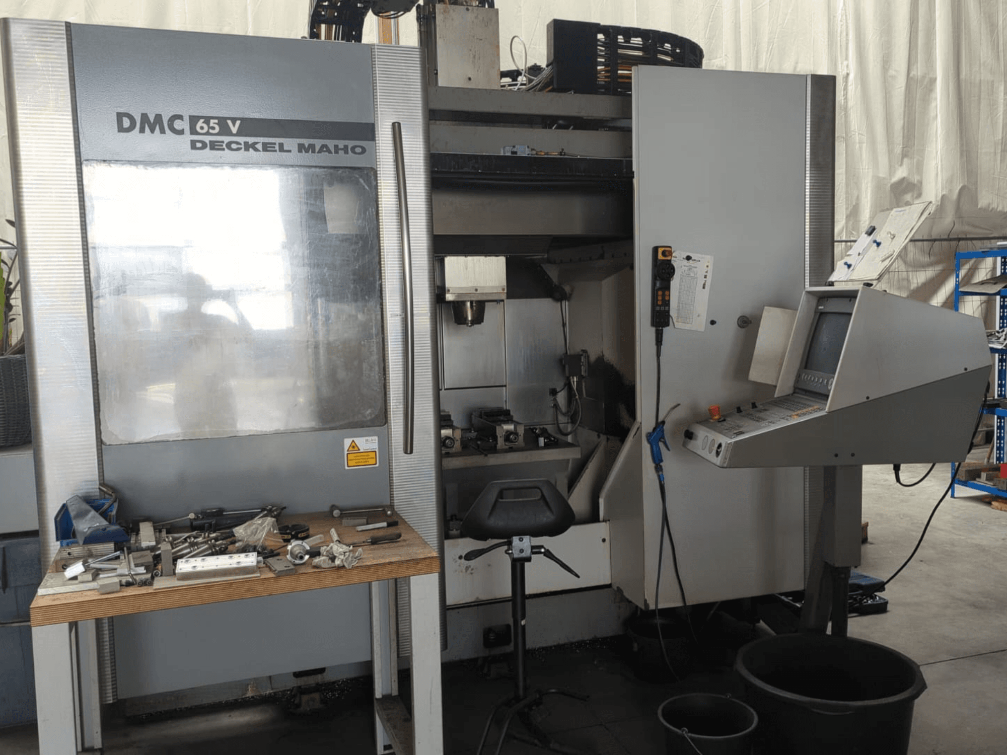 Vooraanzicht van een DECKEL MAHO DMC 65 V verticaal bewerkingscentrum met bedieningspaneel, gereedschappen op een tafel en de inrichting van de werkruimte.