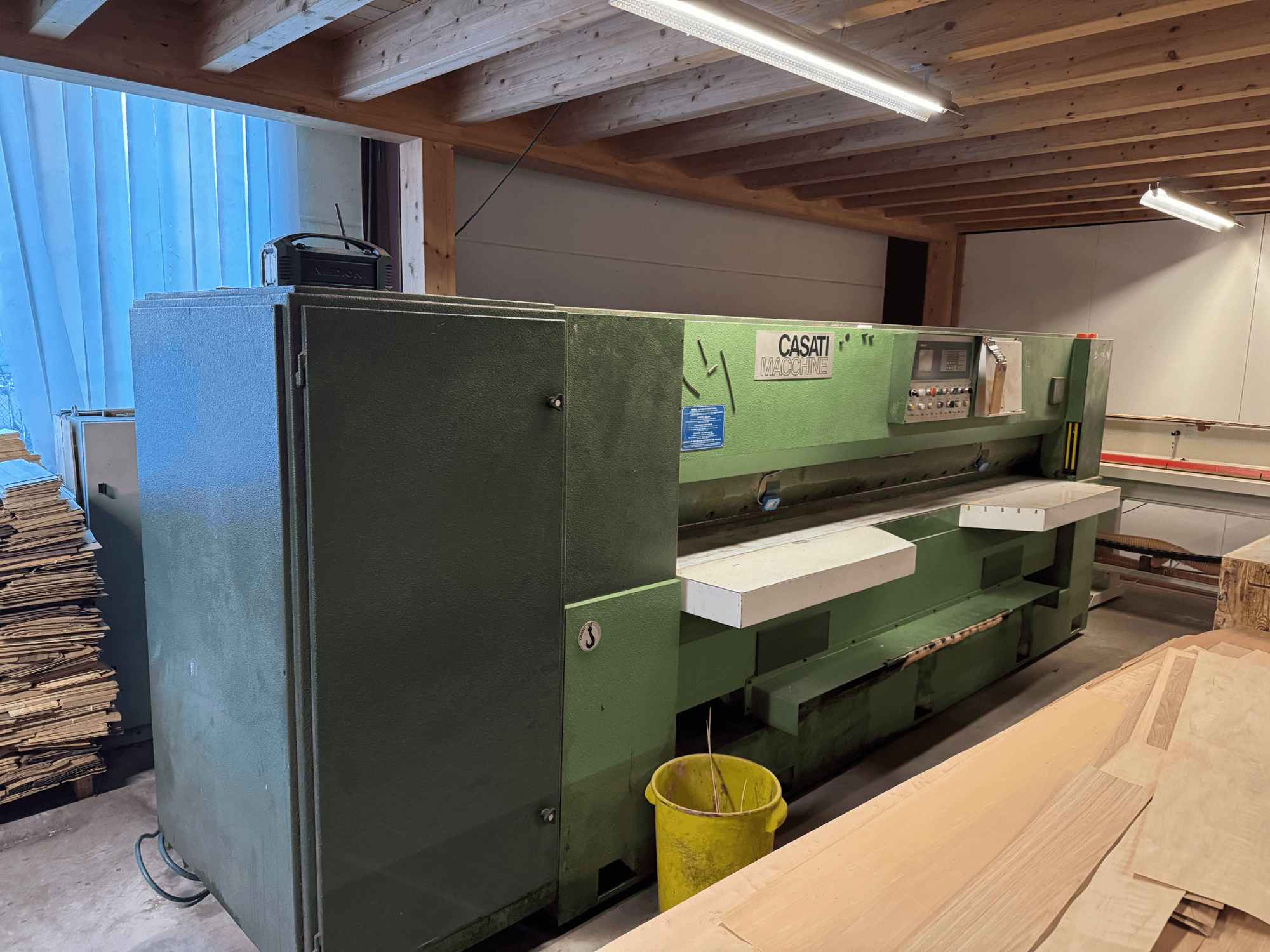 Groene CASATI houtbewerkingsmachine in een werkplaats, vooraanzicht, met bedieningspaneel en witte werkbladen, omringd door houtstapels.