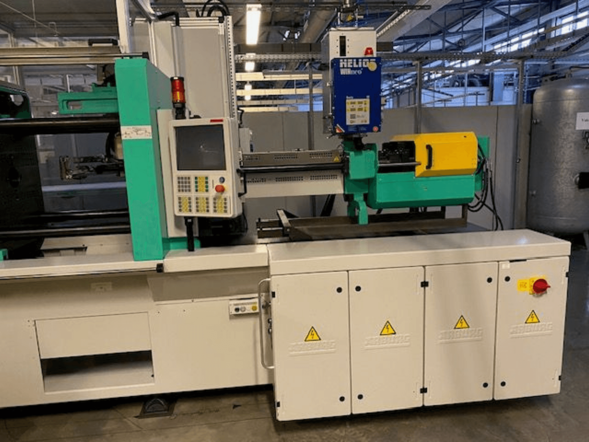 Industriële spuitgietmachine, model HELIO, vooraanzicht, met bedieningspaneel, veiligheidsschermen en noodstopknop.