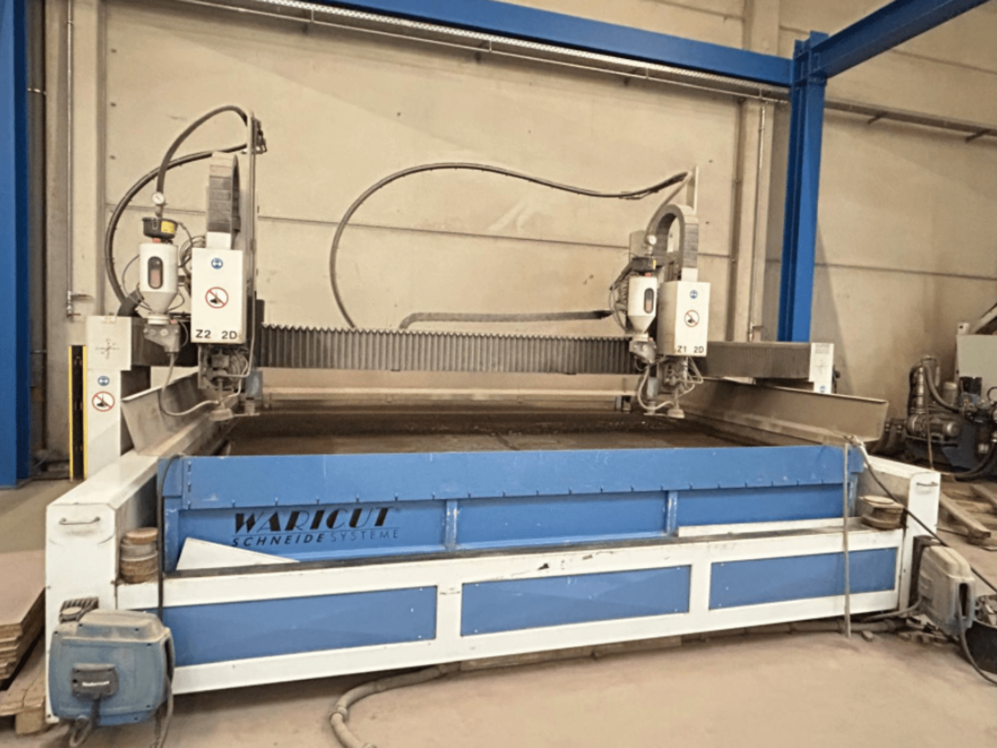 Waterstraalsnijmachine van Waricut, vooraanzicht; met dubbele snijkoppen en blauw werkoppervlak in een werkplaats.