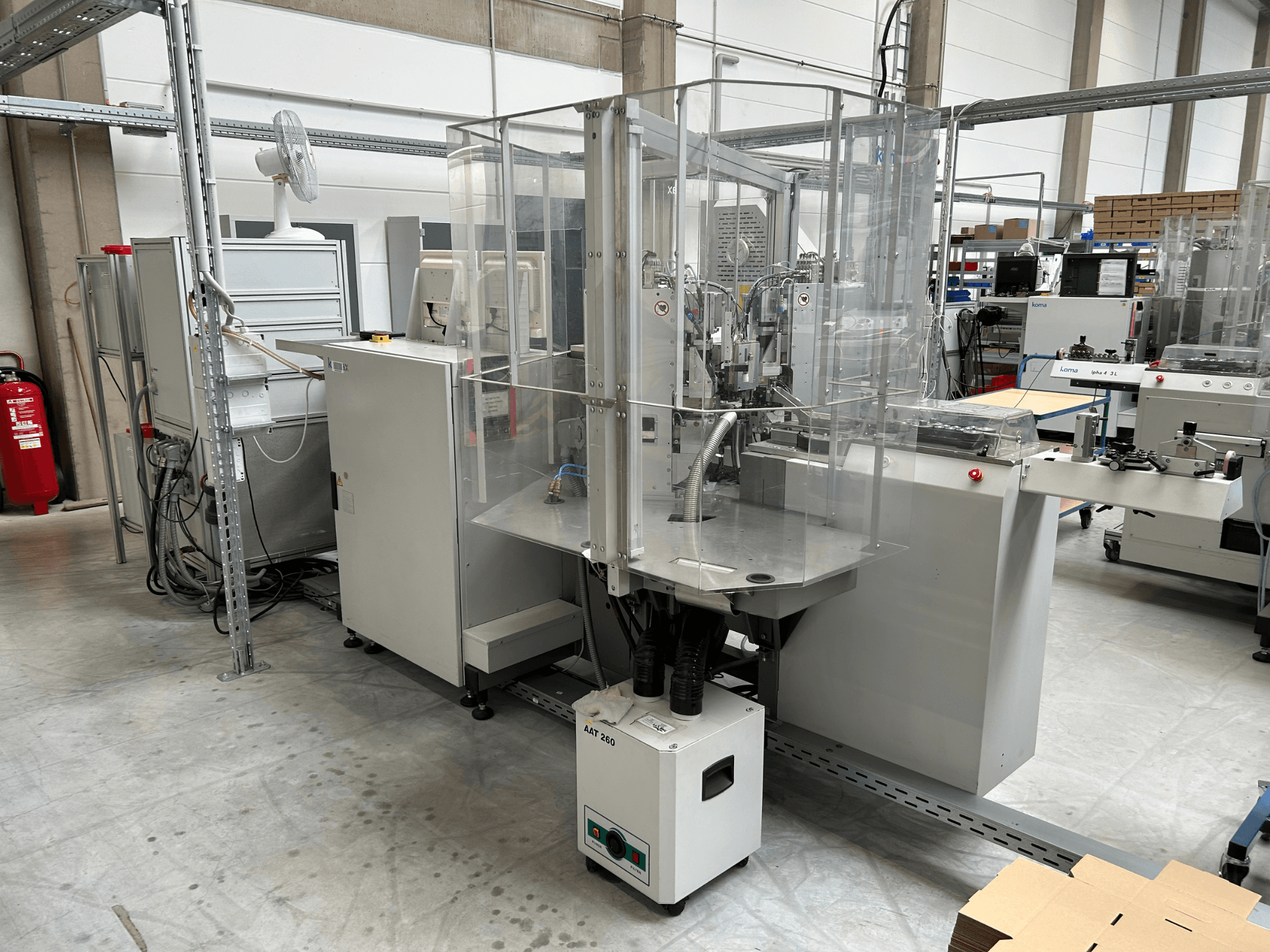 KOMAX Alpha 433 L-automatiseringsapparatuur in een fabrieksomgeving. Voorzien van een doorzichtige veiligheidsbehuizing en diverse bedieningspanelen.