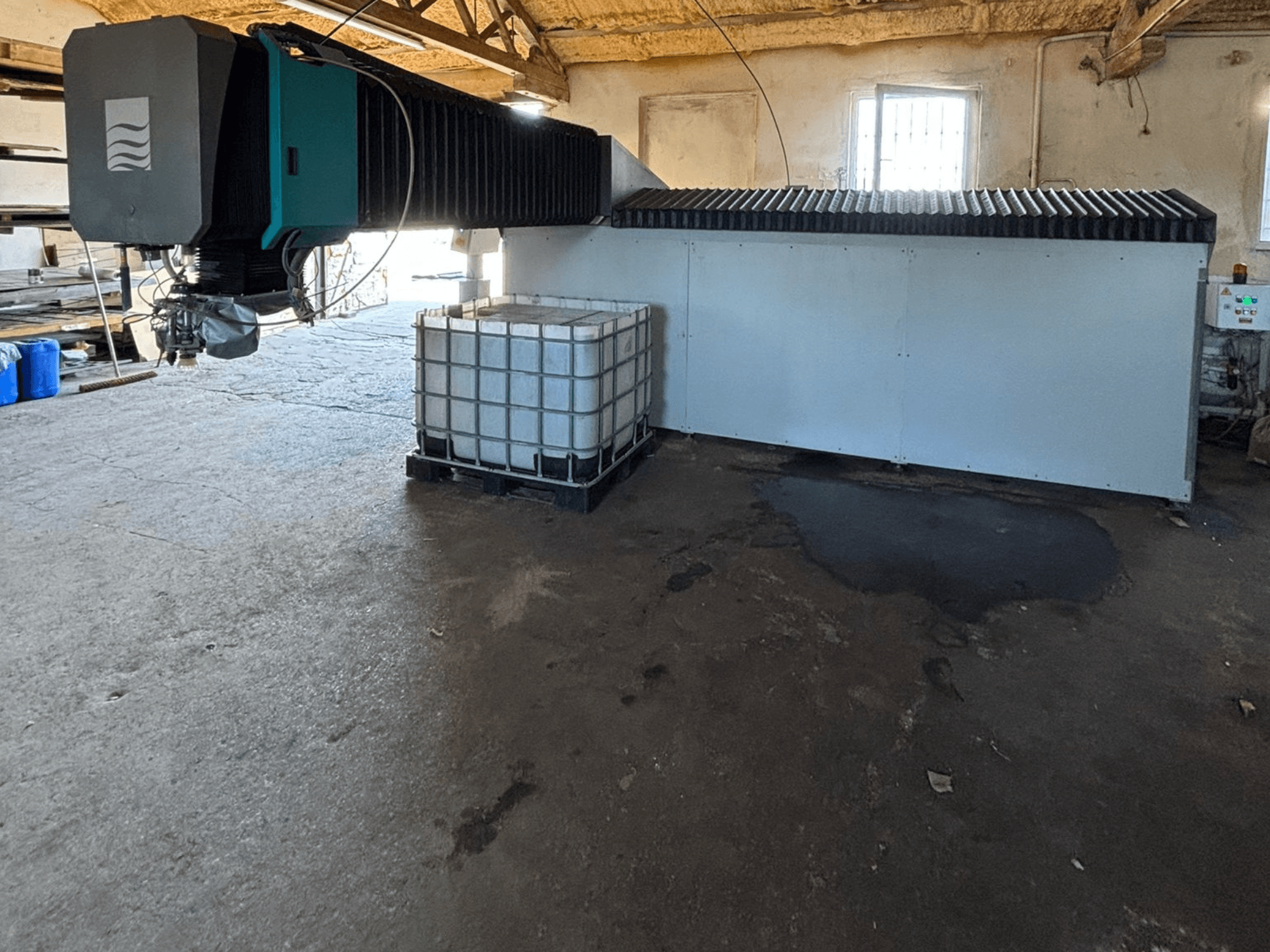 Industriële waterstraalmachine in een werkplaats, zijaanzicht, met een grote watertank en bedieningspaneel ernaast zichtbaar.