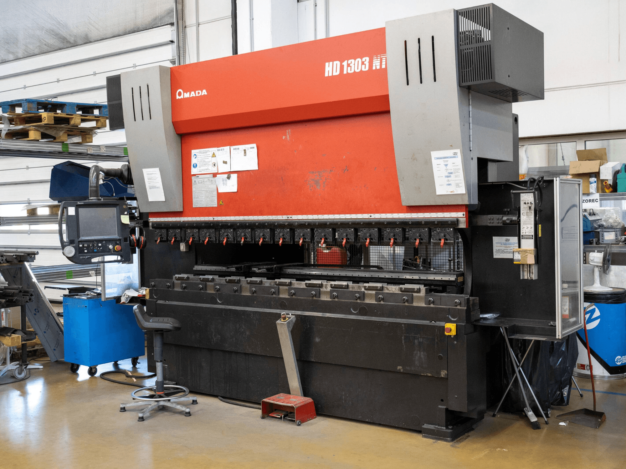 Amada HD 1303 hydraulische afkantpers in een werkplaats, vooraanzicht; voorzien van meerdere klemmen, bedieningspaneel en verstelbare kruk.